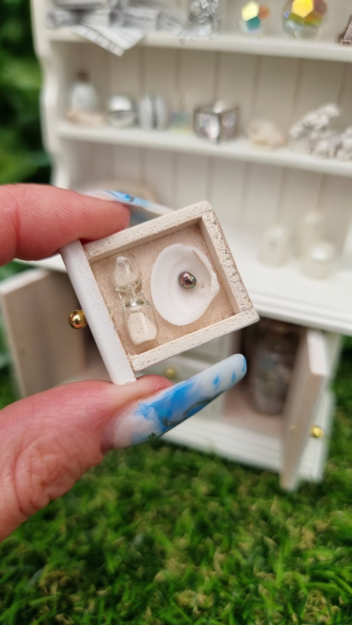Hand holds a miniature 1:12 scale drawer containing a white shell, pearl and tiny sand timer. In the background, a white dresser with counter top and 2 shelves containing candles, crystals, glass bottle, shells, coral and sheet music