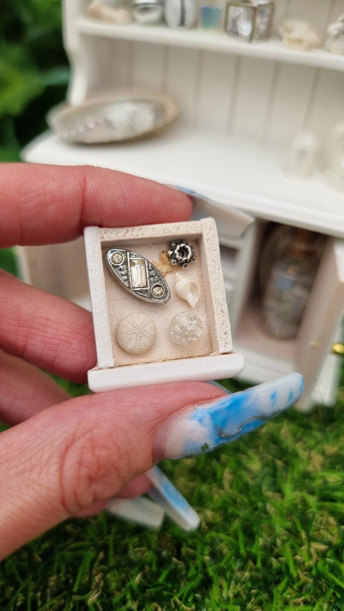 Hand holds a miniature 1:12 scale drawer containing a white shell, sea urchin, leaf, trinket, jewellery box and crackle glass sphere. In the background, a white dresser with counter top and 2 shelves containing candles, crystals, glass bottle, shells, coral and sheet music