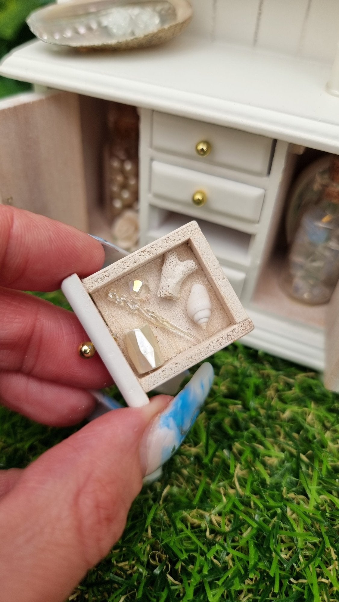 Hand holds a miniature 1:12 scale drawer containing a white shell, crystal, cream box pearlescent horn and coral. In the background, a white dresser with counter top and 2 shelves containing candles, crystals, glass bottle, shells, coral and sheet music