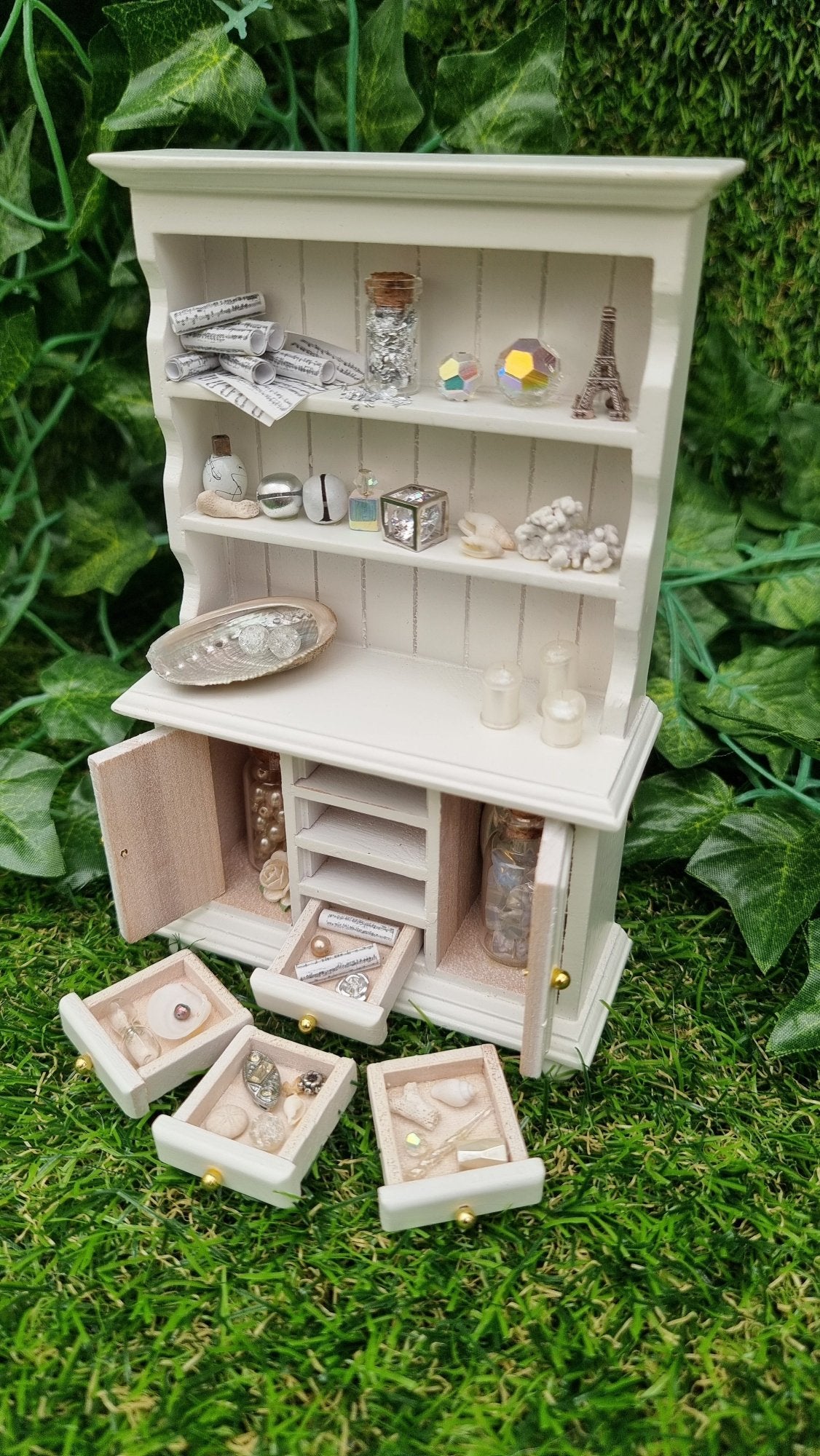 Miniature 1:12 scale white dresser sits against a green background. The dresser has open cupboard doors revealing a white rose and bottles of gemstones. 4 drawers are arranged in front containing light-hued curios and trinkets. The counter top and 2 shelves above contain more candles, crystals, glass bottle, shells, coral and sheet music