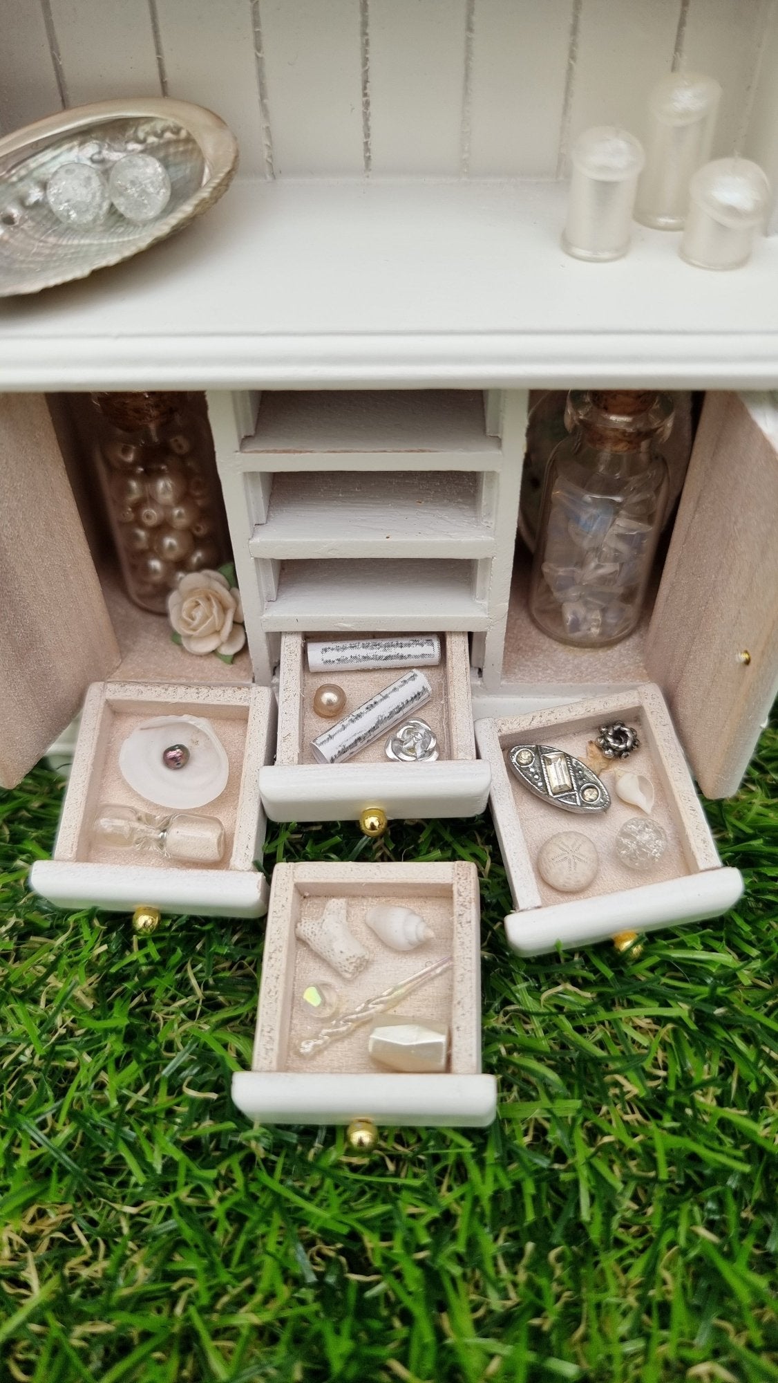 Miniature 1:12 scale white dresser sits against a green background. The dresser has open cupboard doors revealing a white rose and bottles of gemstones. 4 drawers are arranged in front containing light-hued curios and trinkets. The counter top above has candles and a shell with crackle-glass spheres on a shell