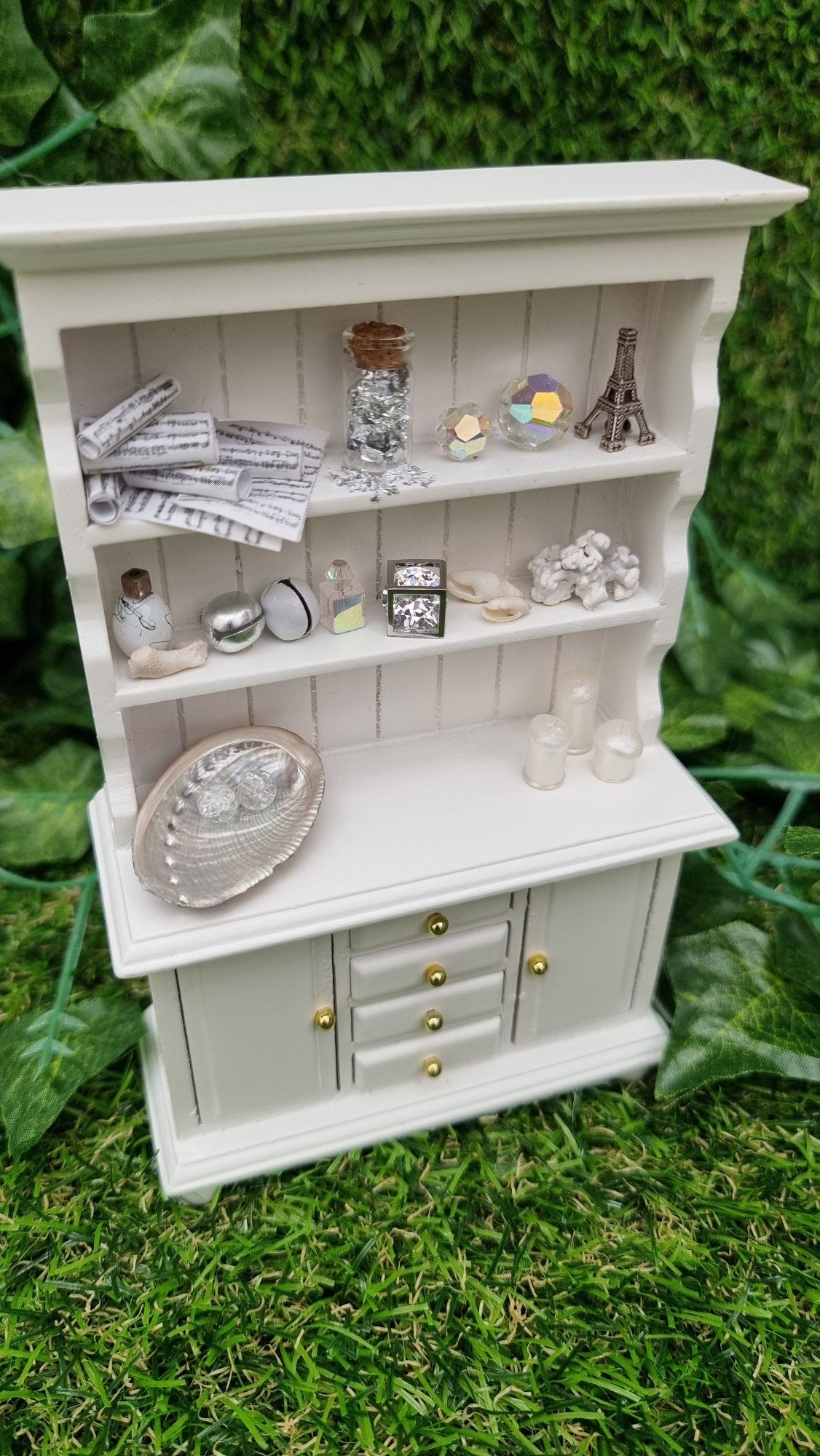 Miniature 1:12 scale white dresser sits against a green background. The dresser has closed cupboard doors and 4 closed drawers. The counter top and 2 shelves above contain candles, crystals, glass bottle, shells, coral and sheet music