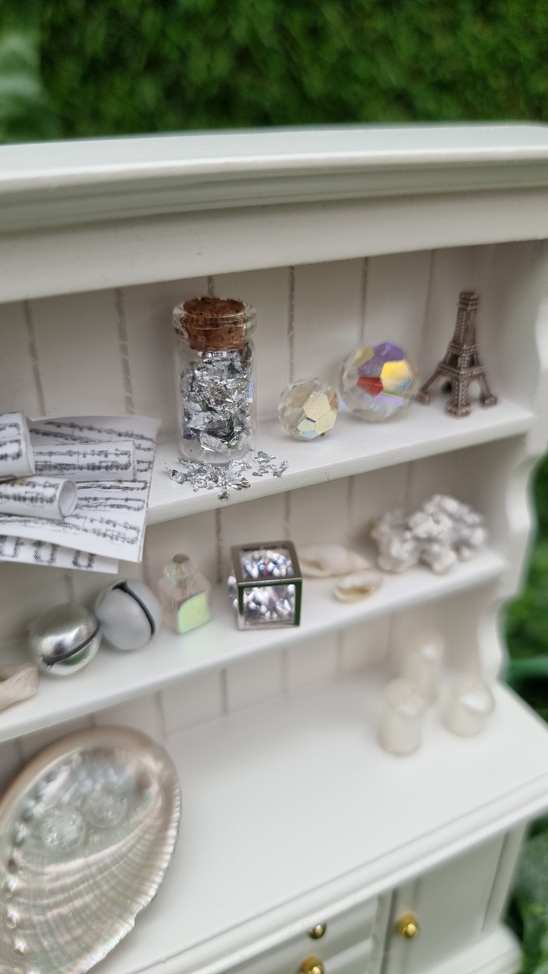 Detail shown of a miniature 1:12 scale white dresser, showing the counter top and 2 shelves displaying candles, crystals, glass bottle, eiffel tower sculpture, bells, vessels, shells, crackle-glass spheres, coral and sheet music. The dreeser sits against a green background