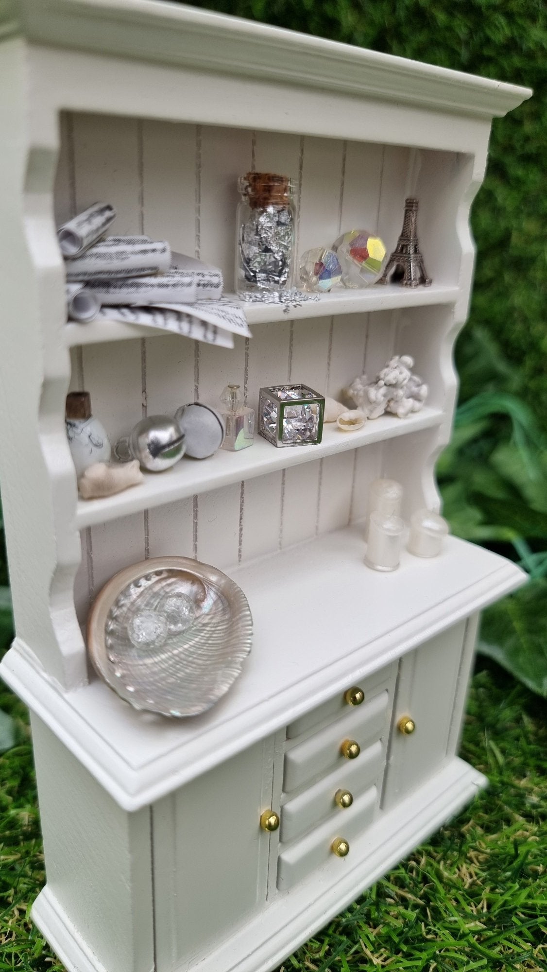 Miniature 1:12 scale white dresser sits against a green background. The dresser has closed cupboard doors and 4 closed drawers. The counter top and 2 shelves above contain candles, crystals, glass bottle, shells, coral and sheet music