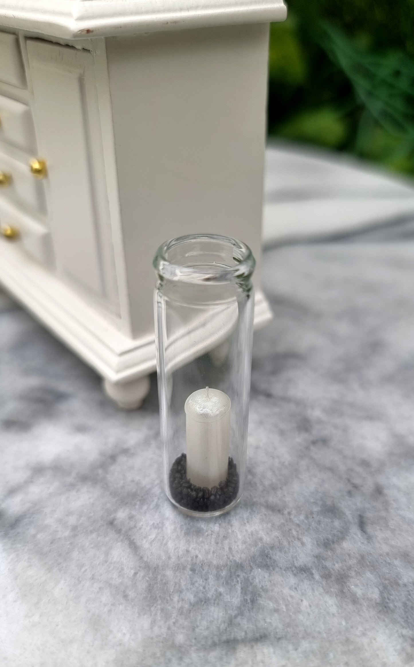 A tall 1:12 scale glass hurricane lantern containing a pearlescent white candle and black sand sits beside a miniature white dresser.