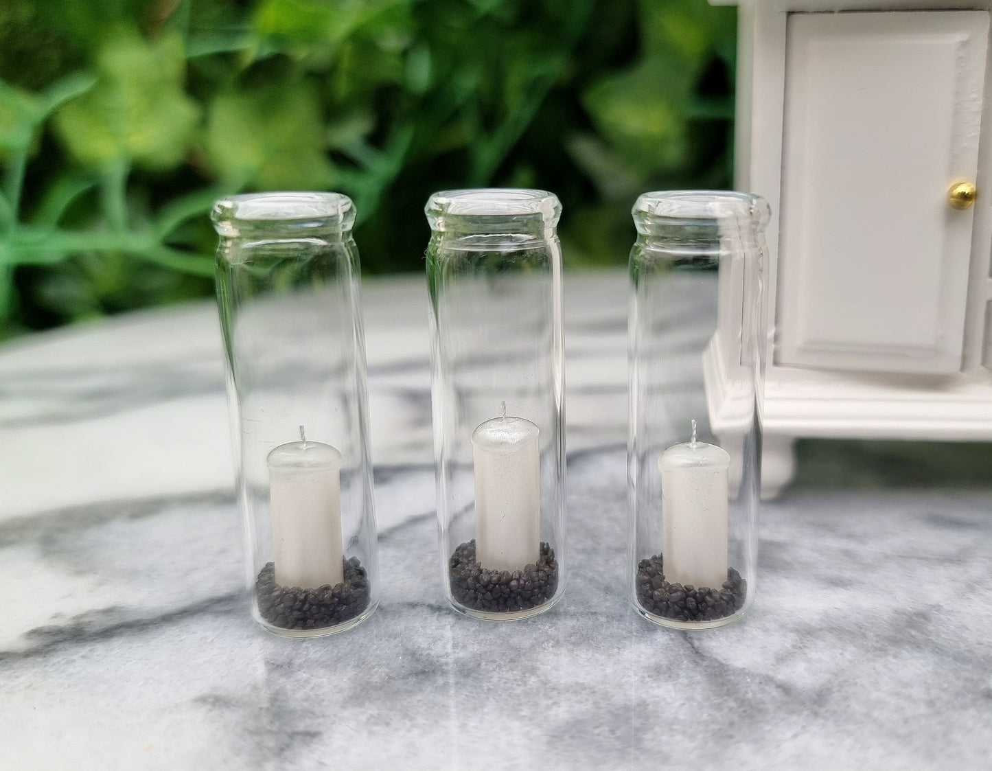 A row of 3 tall 1:12 scale glass hurricane lanterns containing a pearlescent white candles and black sand sit beside a miniature white dresser.