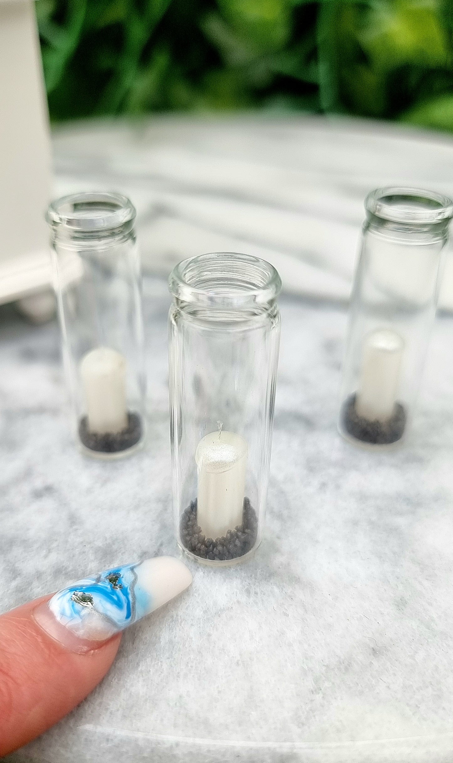 A finger points at a tall 1:12 scale glass hurricane lantern containing a pearlescent white candle and black sand. A miniature white dresser sits in the background