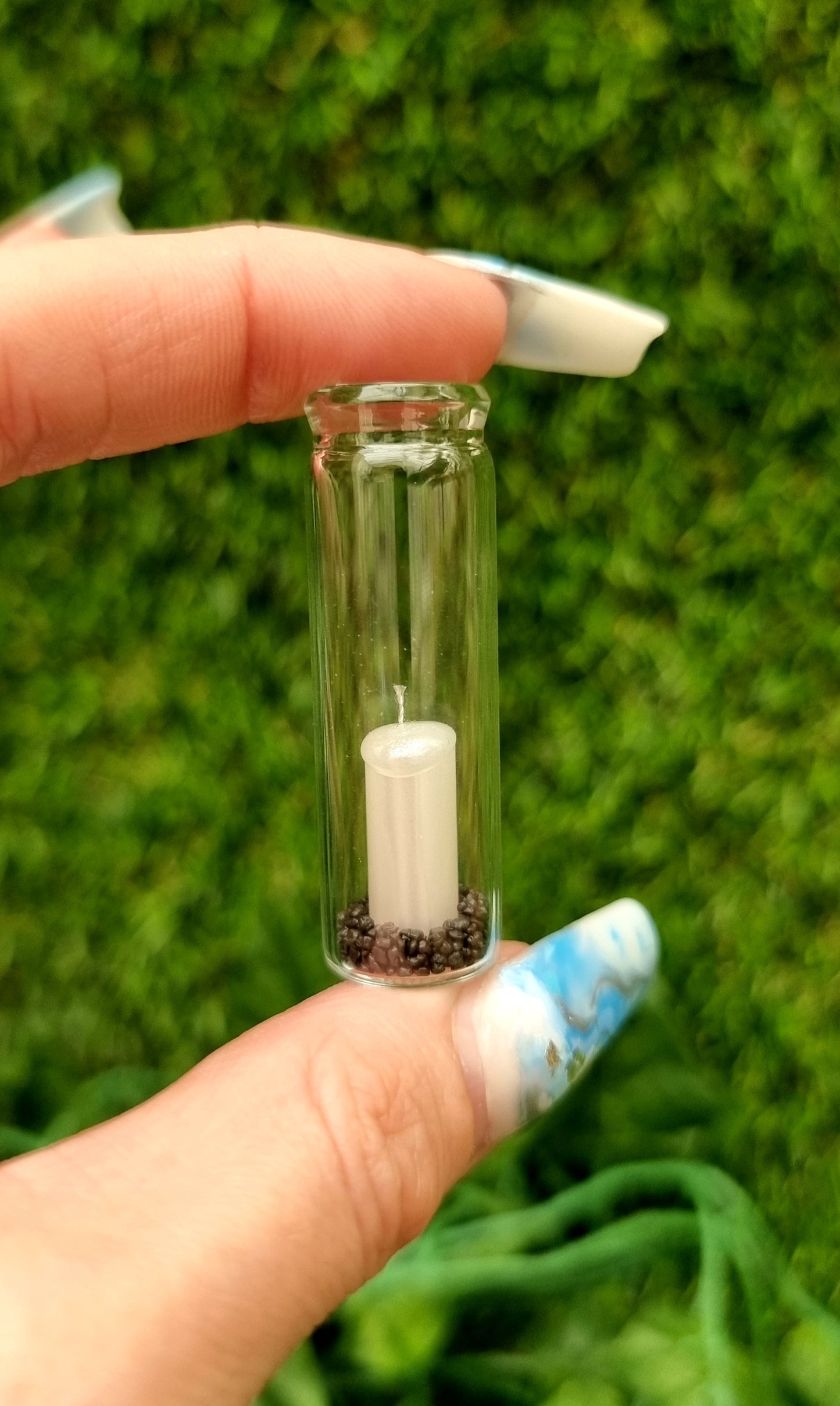 A tall 1:12 scale glass hurricane lantern containing a pearlescent white candle and black sand is held between 2 fingers against a green background