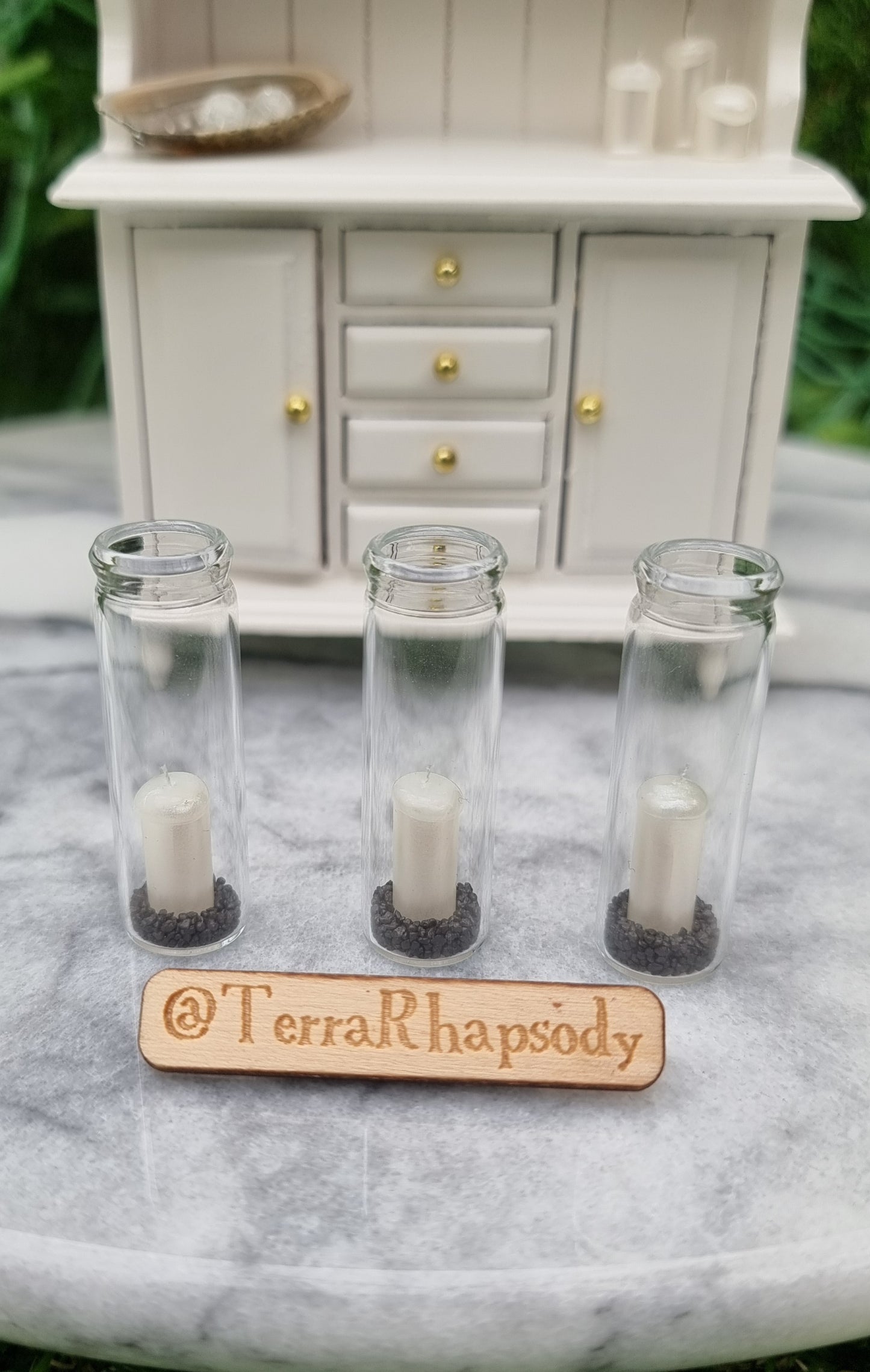 A row of 3 tall 1:12 scale glass hurricane lanterns containing a pearlescent white candles and black sand sit in front of a miniature white dresser. @TerraRhapsody is written on a piece of wood in front.