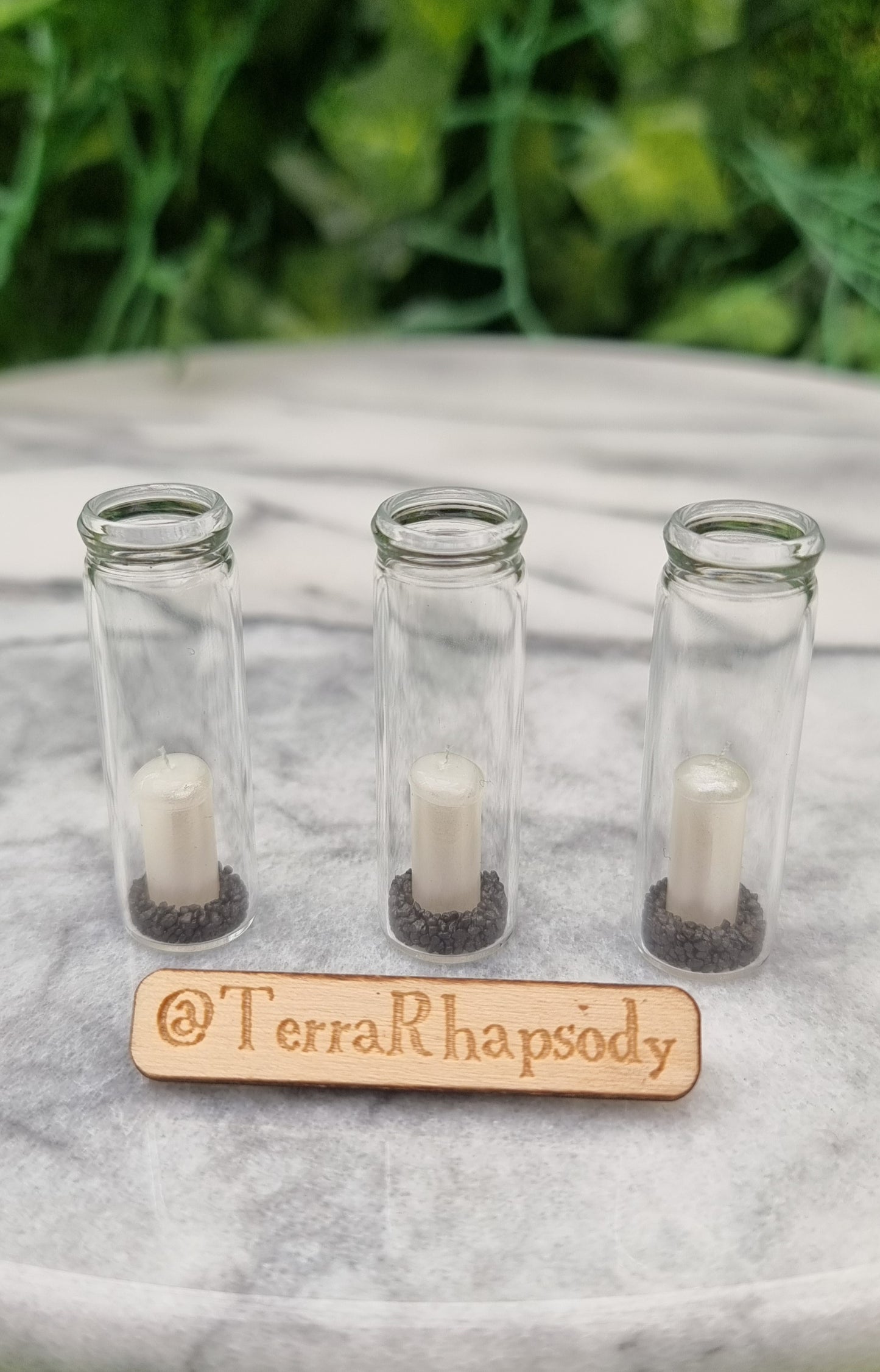 A row of 3 tall 1:12 scale glass hurricane lanterns containing a pearlescent white candles and black sand sit on a piece of marble. @TerraRhapsody is written on a piece of wood in front.