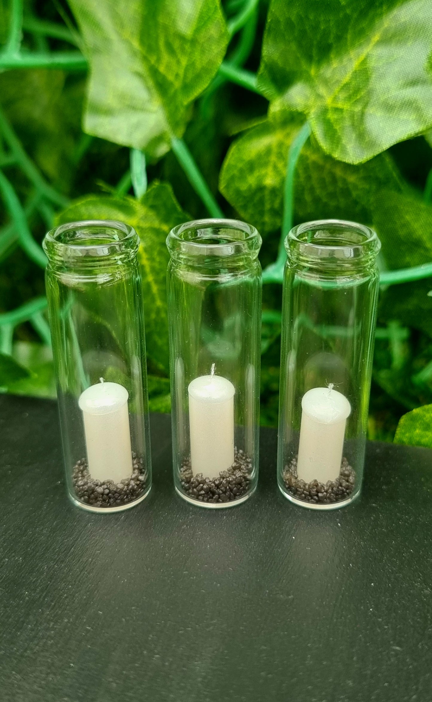 A row of 3 tall 1:12 scale glass hurricane lanterns containing a pearlescent white candles and black sand sit on a black base in front of green background