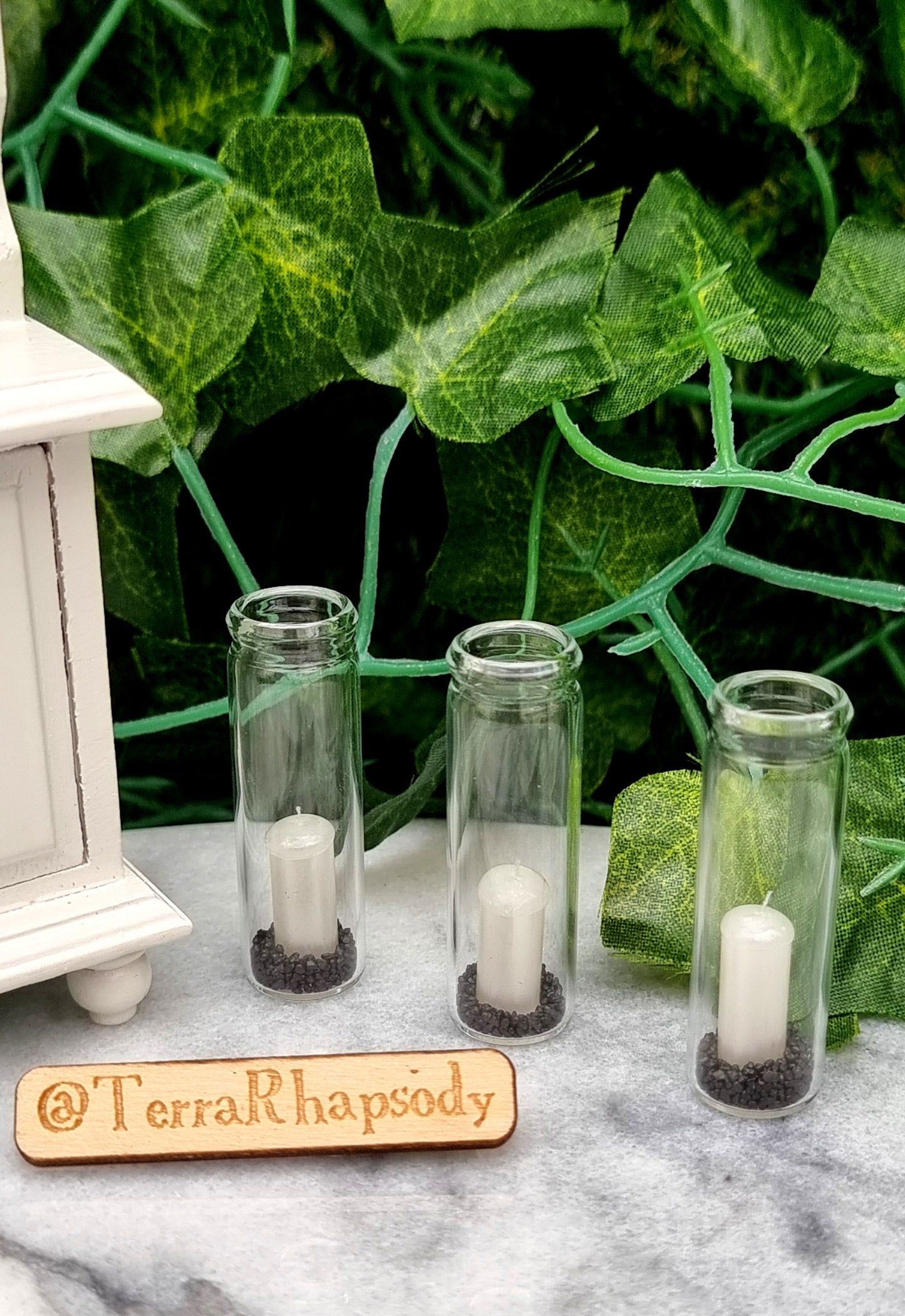A row of 3 tall 1:12 scale glass hurricane lanterns containing a pearlescent white candles and black sand sit beside a miniature white dresser. @TerraRhapsody is written on a piece of wood in front.