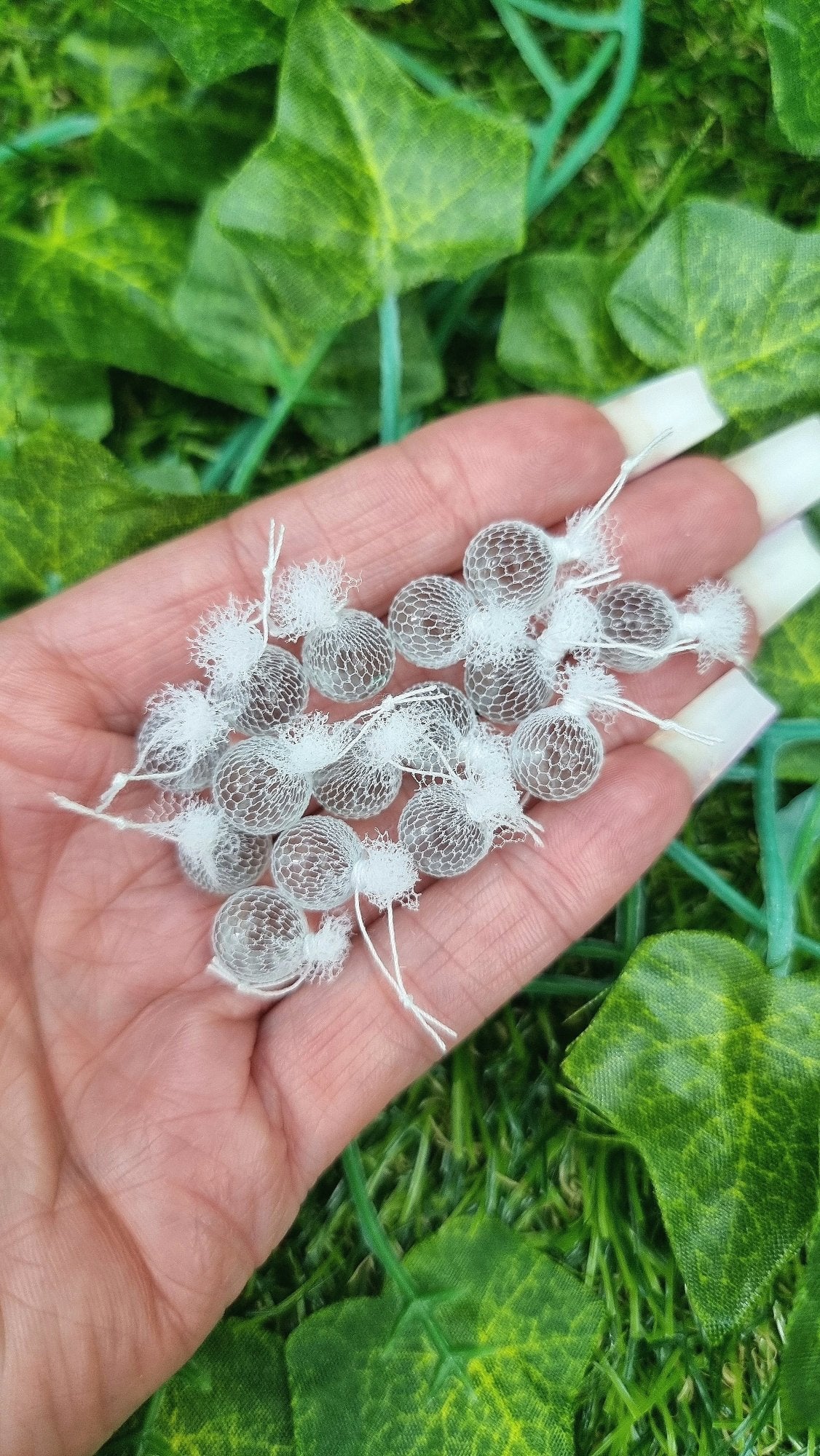 A hand holds 15 miniature 1:12 scale clear glass fishing floats covered in white netting with a loop of white rope at the top of each. Held against green background