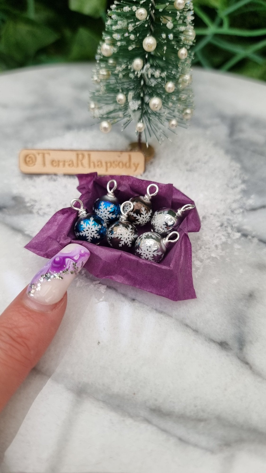 Miniature 1:12 scale open tray box contains 6 Christmas baubles. The baubles are silver, grey and blue with white snowflake motifs. The box is lined with purple paper, displayed on snow. @TerraRhapsody is written on wood in the background
