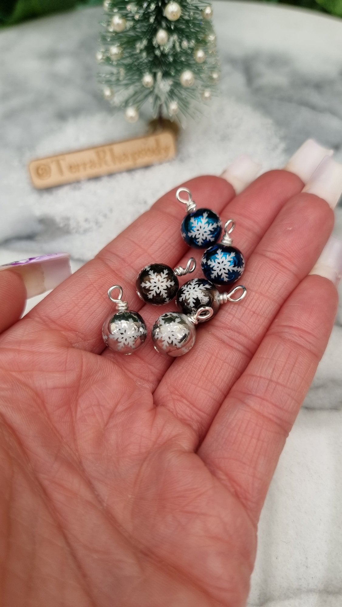 6 Miniature 1:12 Christmas baubles are displayed on snow in front of a Christmas tree. The baubles are silver, grey and blue with white snowflake motifs. @TerraRhapsody is written on wood in the background