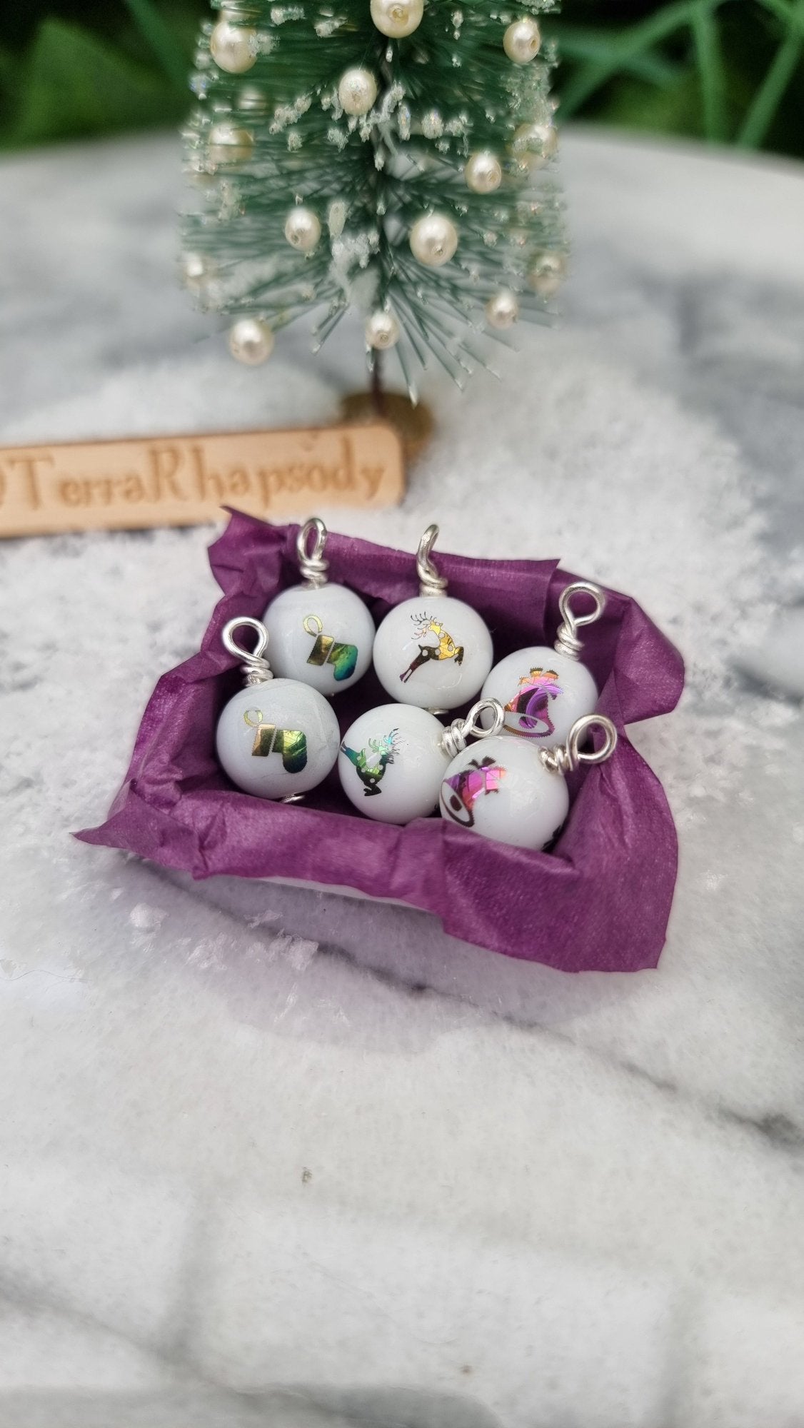 Miniature 1:12 scale open tray box contains 6 Christmas baubles. The baubles are white with multicoloured festive motifs. The box is lined with purple paper, displayed on snow and marble. @TerraRhapsody is written on wood in the background