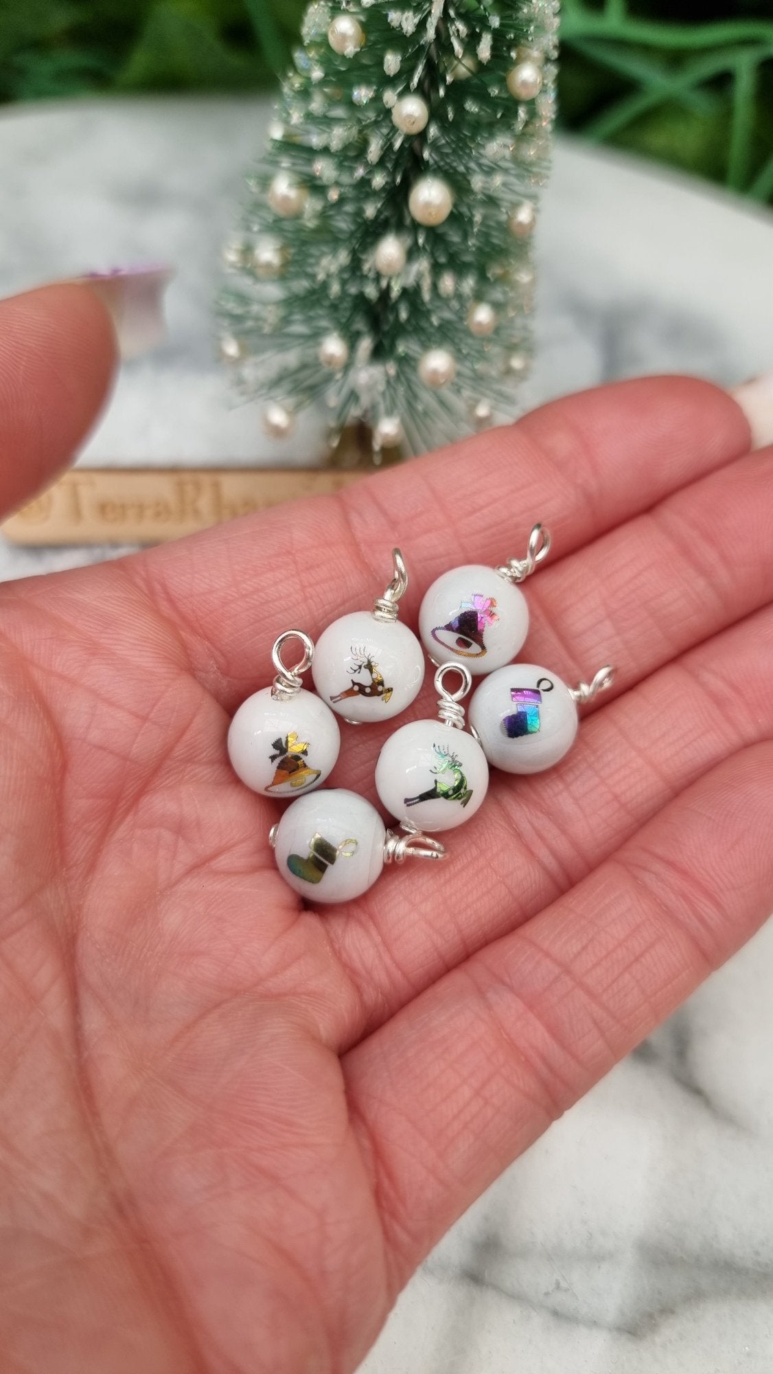 6 Miniature 1:12 Christmas baubles are displayed in an open hand. The baubles are white with multicoloured christmas motifs. @TerraRhapsody is written on wood in the background