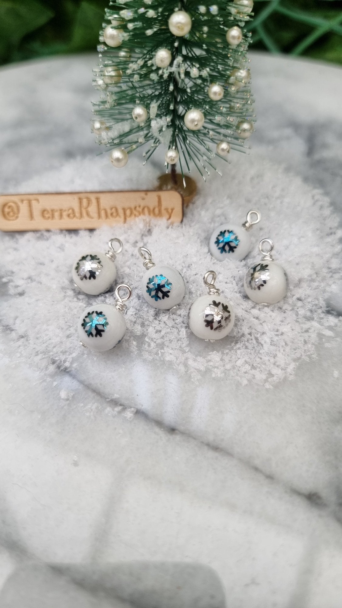 6 Miniature 1:12 Christmas baubles are displayed in an open hand. The baubles are white with silver and blue snowflake motifs. @TerraRhapsody is written on wood in the background