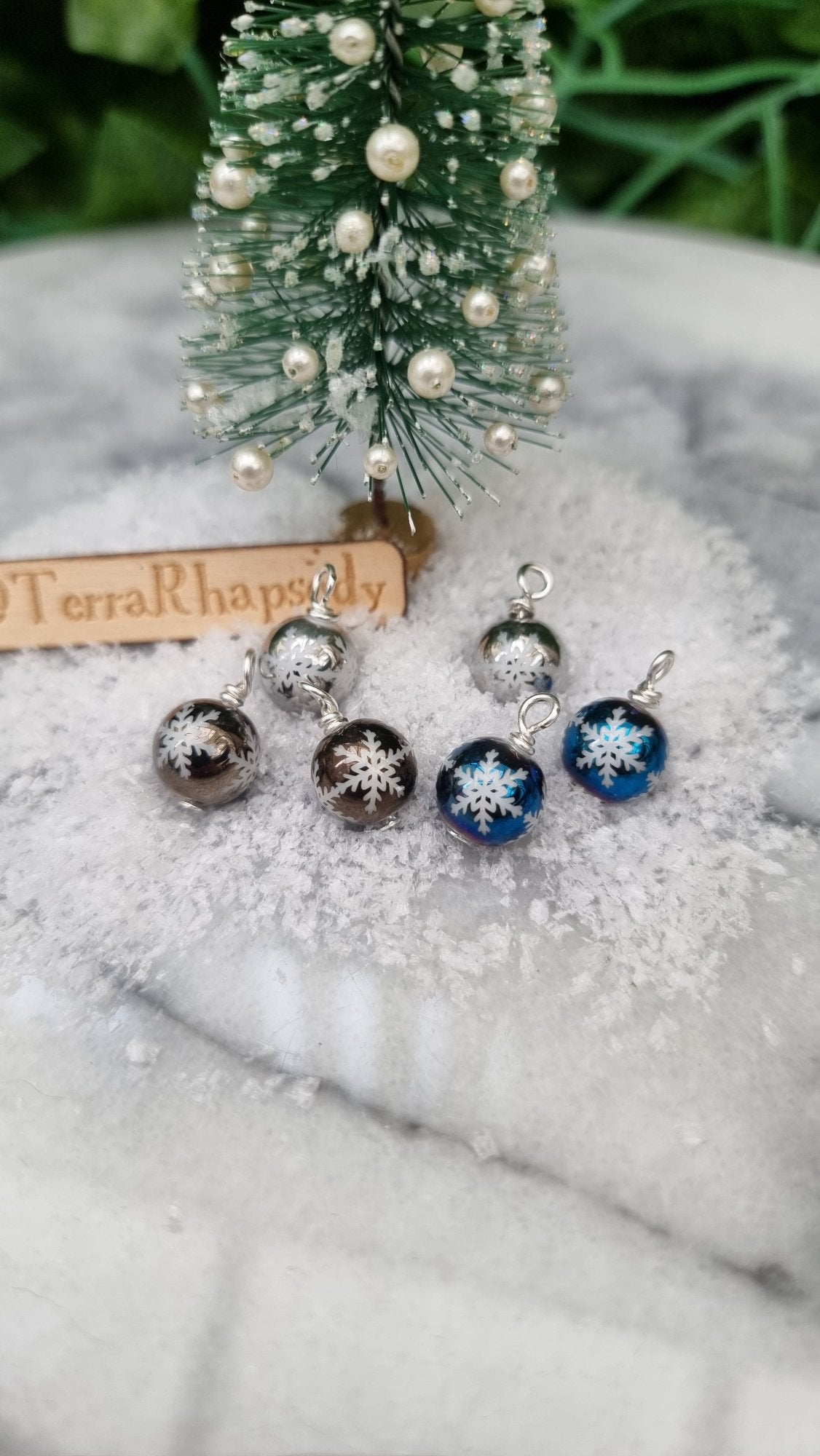 6 Miniature 1:12 Christmas baubles are displayed on snow in front of a Christmas tree. The baubles are silver, grey and blue with white snowflake motifs. @TerraRhapsody is written on wood in the background