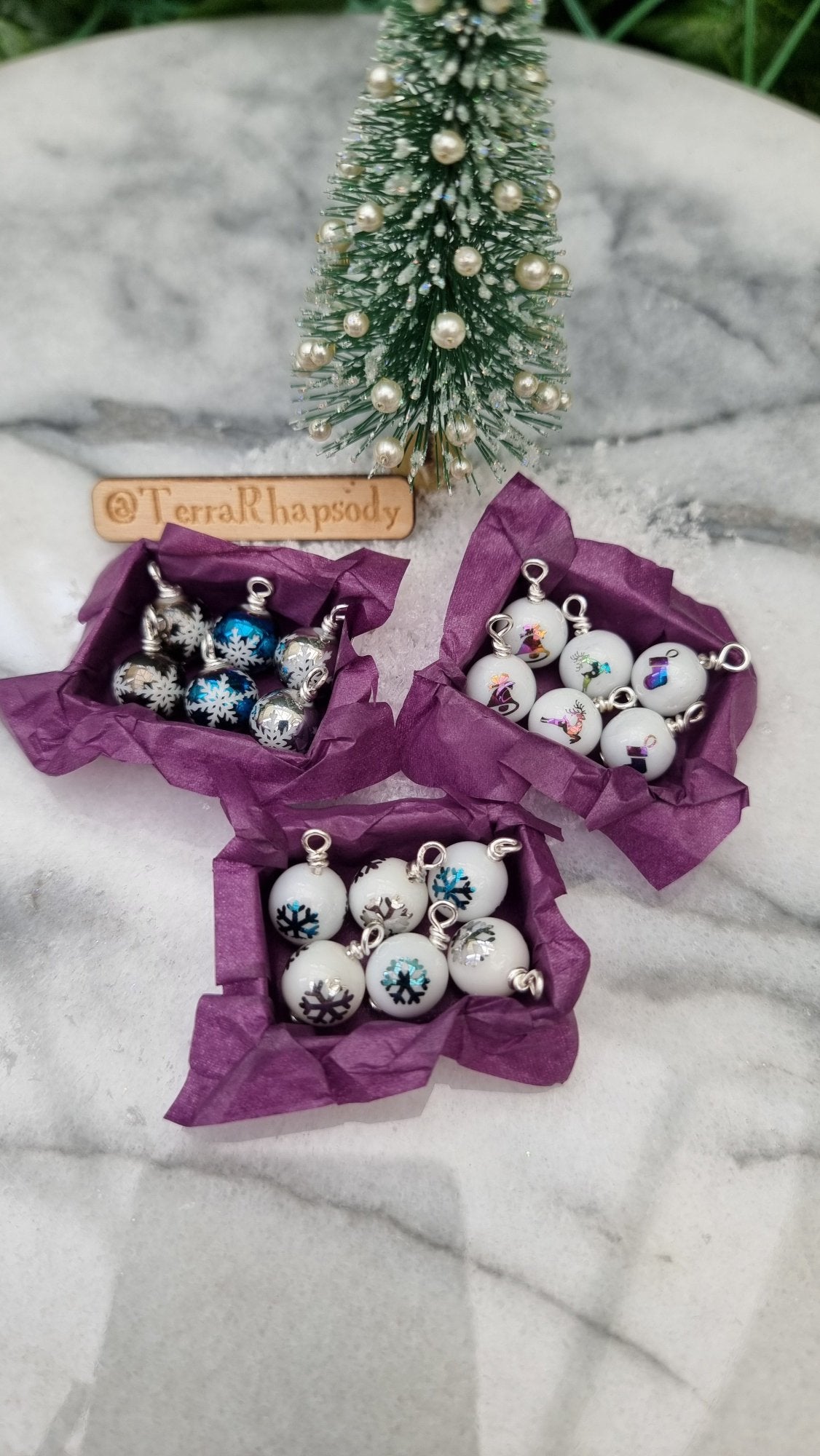 3 miniature 1:12 scale open tray boxes contain 6 Christmas baubles. The baubles are white with coloured festive motifs. The boxes are lined with purple paper displayed in front of a Christmas tree. @TerraRhapsody is written on wood in the background