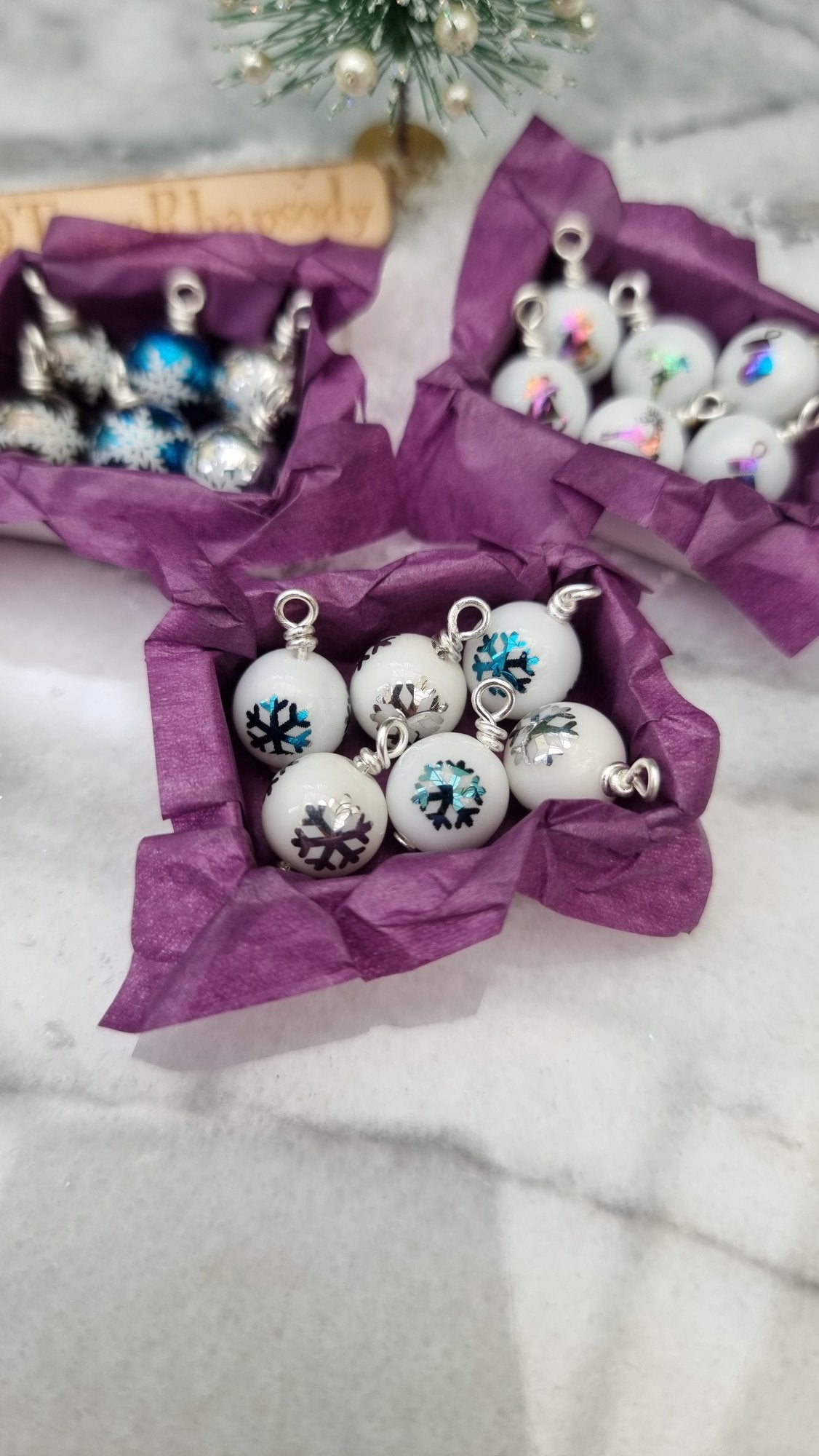 3 miniature 1:12 scale open tray boxes contain 6 Christmas baubles. The baubles are white with coloured festive motifs. The boxes are lined with purple paper displayed in front of a Christmas tree. @TerraRhapsody is written on wood in the background