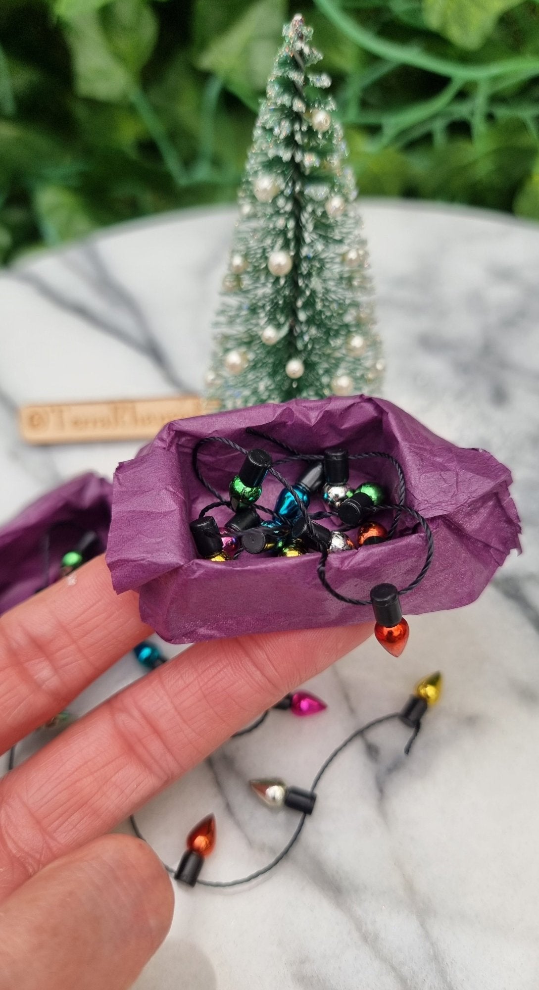 Hand holds a 1:12 scale miniature box of Christmas lights. The box is lined with purple paper filled with multicoloured lights on black string. @TerraRhapsody is written on wood behind it