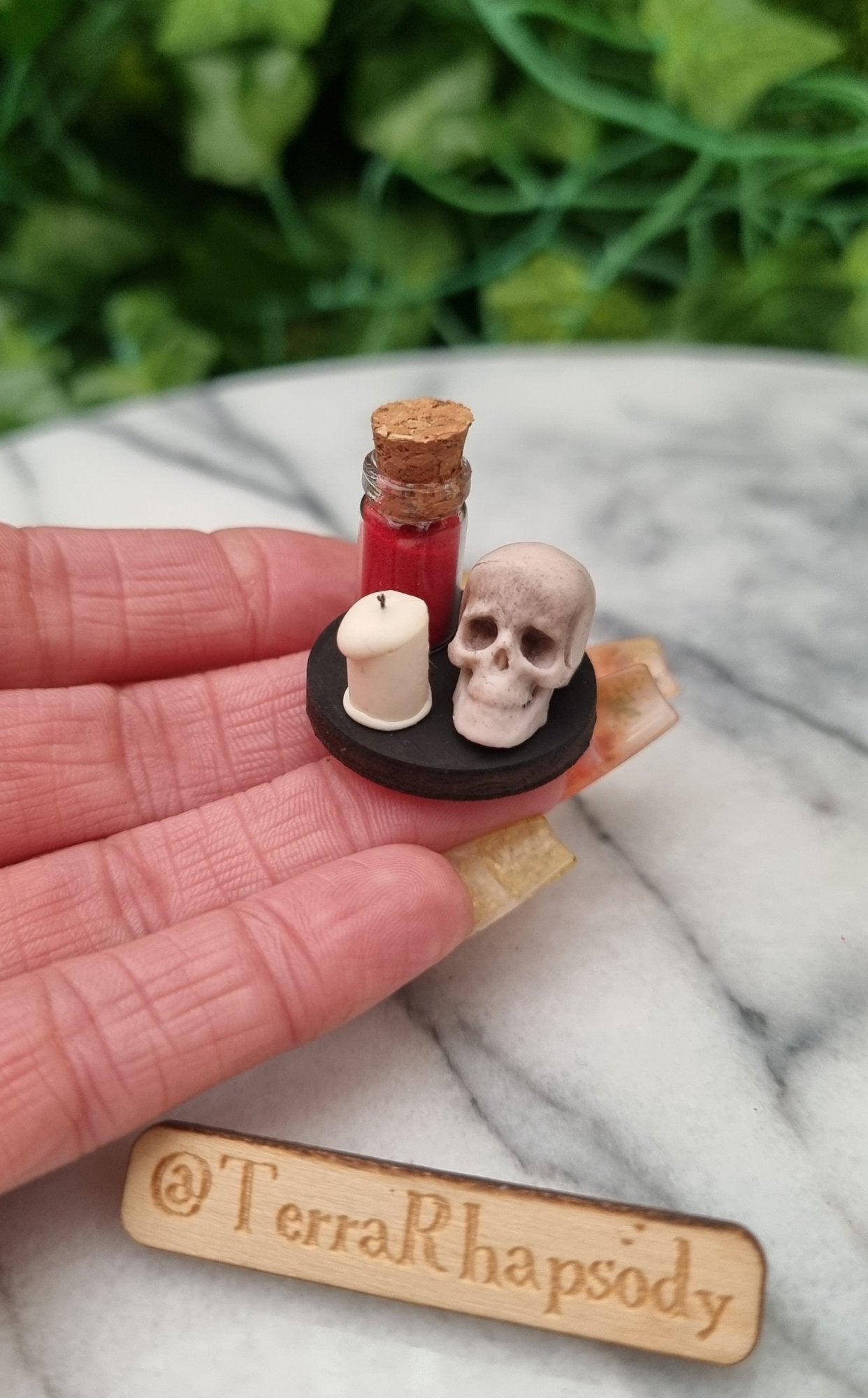 1:12 scale miniature Halloween tray sits on a hand. The tray is black with a detailed skull, ivory candle with black wick and a glass bottle with red powder inside. @TerraRhapsody is written on wood in the foreground