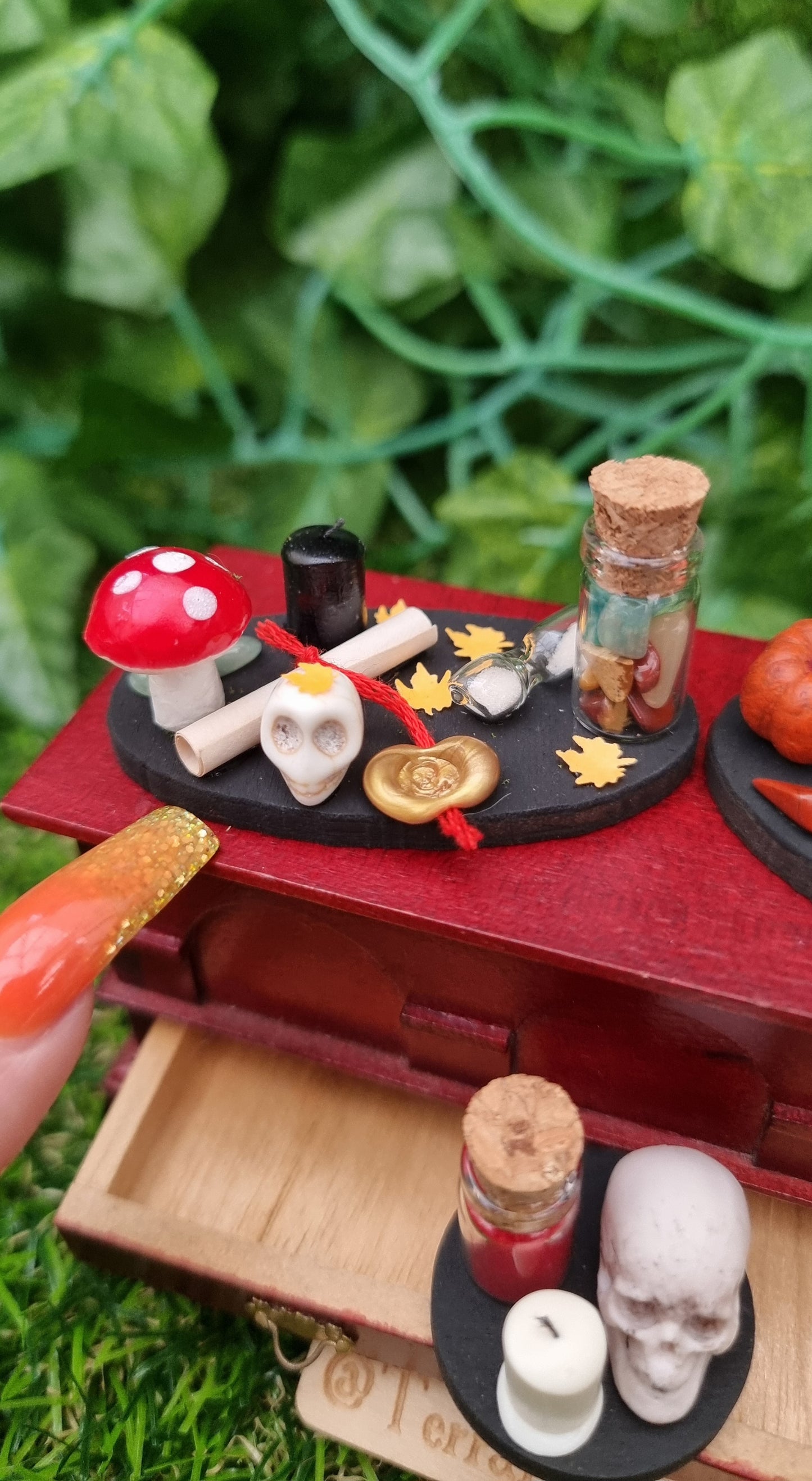 1:12 scale black tray displaying Halloween and Autumn/fall items sat on miniature chest. The tray is oval with fly agaric mushroom, bottle of gemstones, paper scroll with gold seal, skull, black candle, orange leaves and sand timer. Afinger pointe for scale and @TerraRhapsody is written on wood in the foreground
