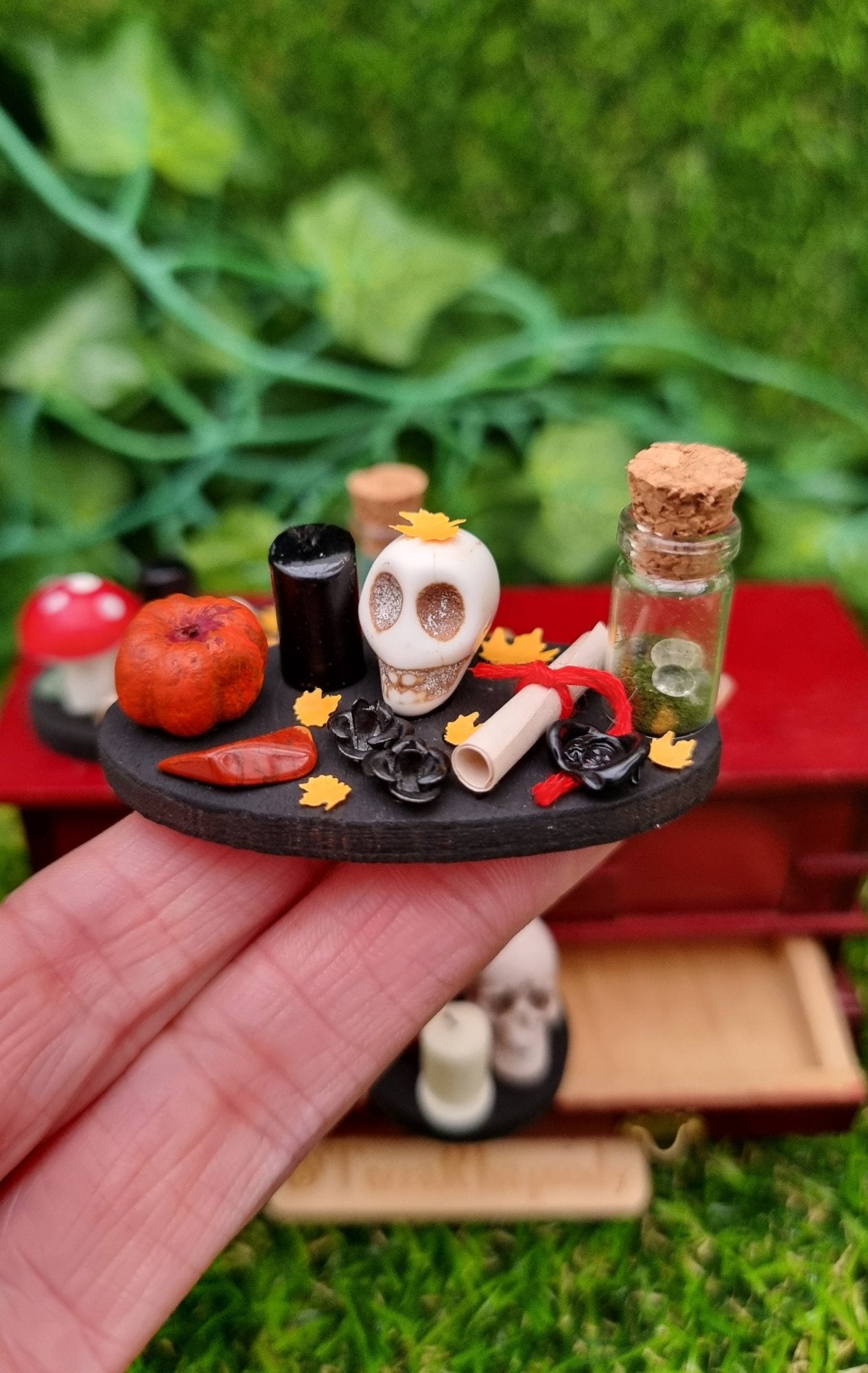 1:12 scale black tray displaying Halloween and Autumn/fall items sat on a hand. The tray is oval with pumpkin, bottle of green foliage and clear spheres, paper scroll with black seal, skull, black candle, orange leaves, black roses and gemstone. @TerraRhapsody is written on wood