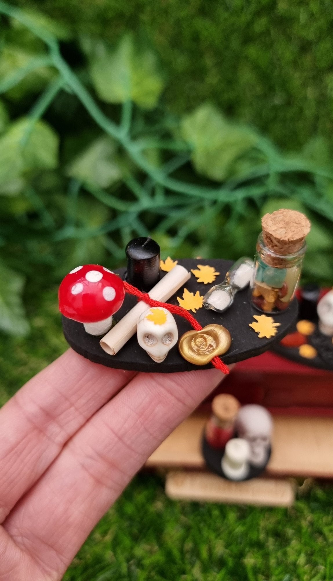 1:12 scale black tray displaying Halloween and Autumn/fall items sat on a hand. The tray is oval with fly agaric mushroom, bottle of gemstones, paper scroll with gold seal, skull, black candle, orange leaves and sand timer. @TerraRhapsody is written on wood