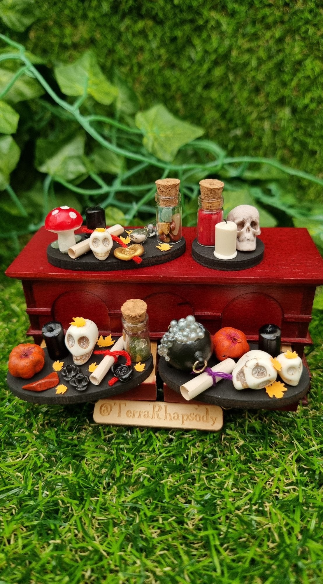 4 1:12 scale black trays displaying Halloween and Autumn/fall items sat on a mini chest. 1 tray is round with skull, candle and bottle. 3 trays are oval with pumpkins, glass bottles, paper scrolls, skulls, candles, orange leaves and cauldron.