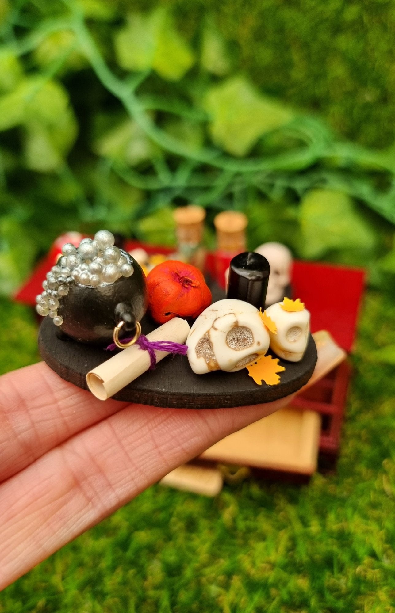 1:12 scale black tray displaying Halloween and Autumn/fall items sat on a hand. The tray is oval with pumpkin, black candle, paper scroll,, a small and large skull, orange leaves and bubbling cauldron. @TerraRhapsody is written on wood