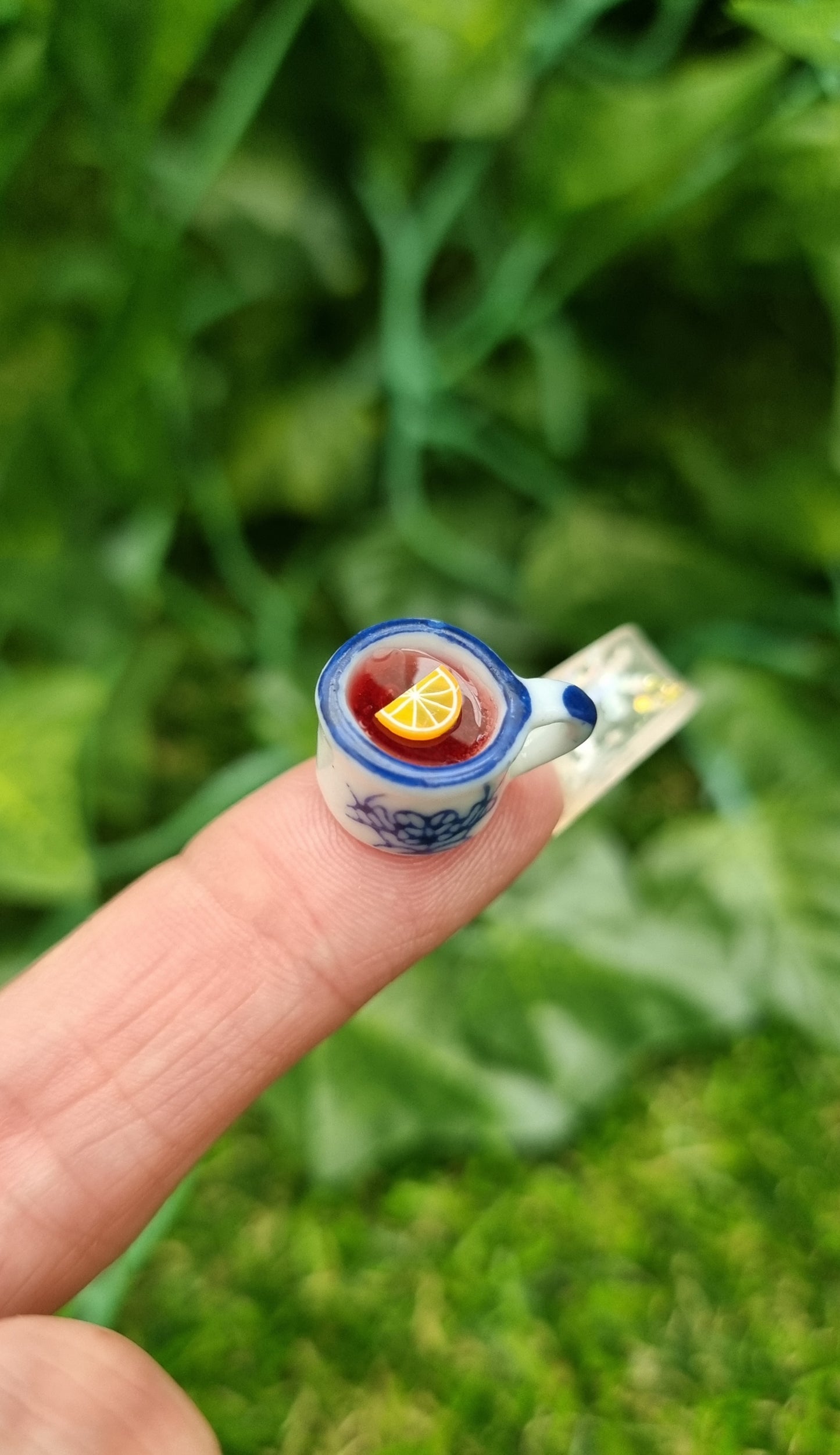 A 1:12 scale miniature mug is held on the end of a long finger. The tiny white and blue porcelain mug contains red liquid (mulled wine), with a slice of orange floating on the surface. The mug has an ornate pattern. Shown against a green background
