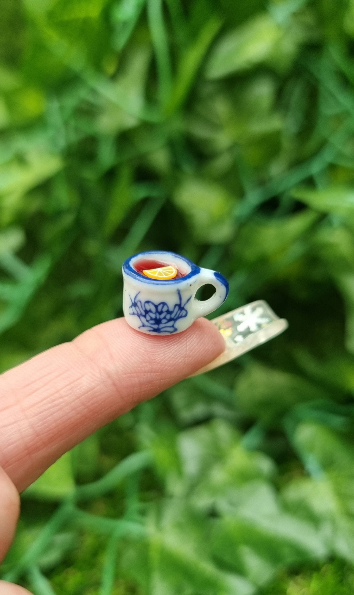 A 1:12 scale miniature mug is held on the end of a finger. The tiny white and blue porcelain mug contains red liquid (mulled wine), with a slice of orange floating on the surface. The mug has an ornate pattern. Shown against a green background