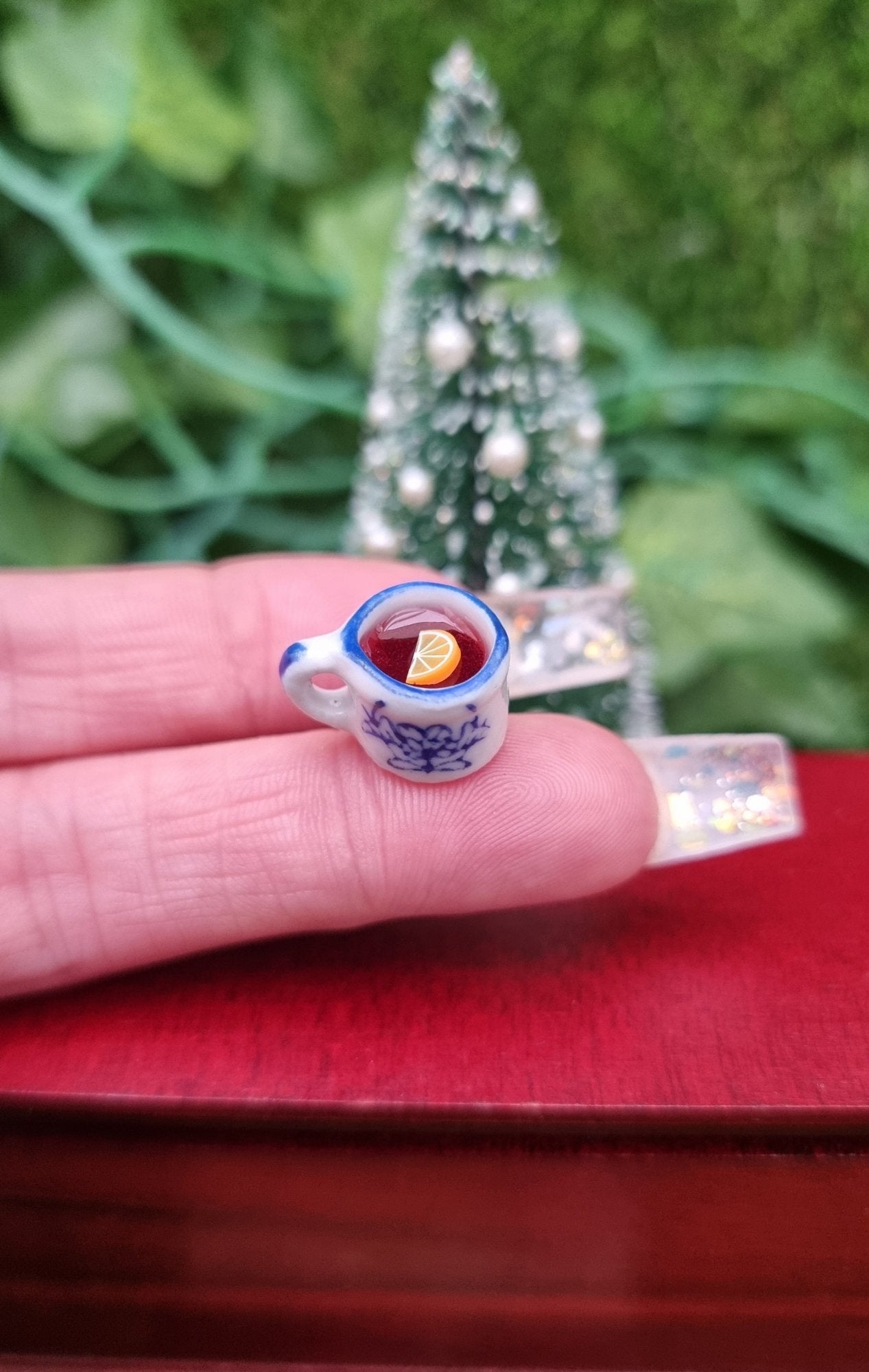 A 1:12 scale miniature mug is held on the end of a finger. The tiny white and blue porcelain mug contains red liquid (mulled wine), with a slice of orange floating on the surface. The mug has an ornate pattern. Shown against a Christmas tree