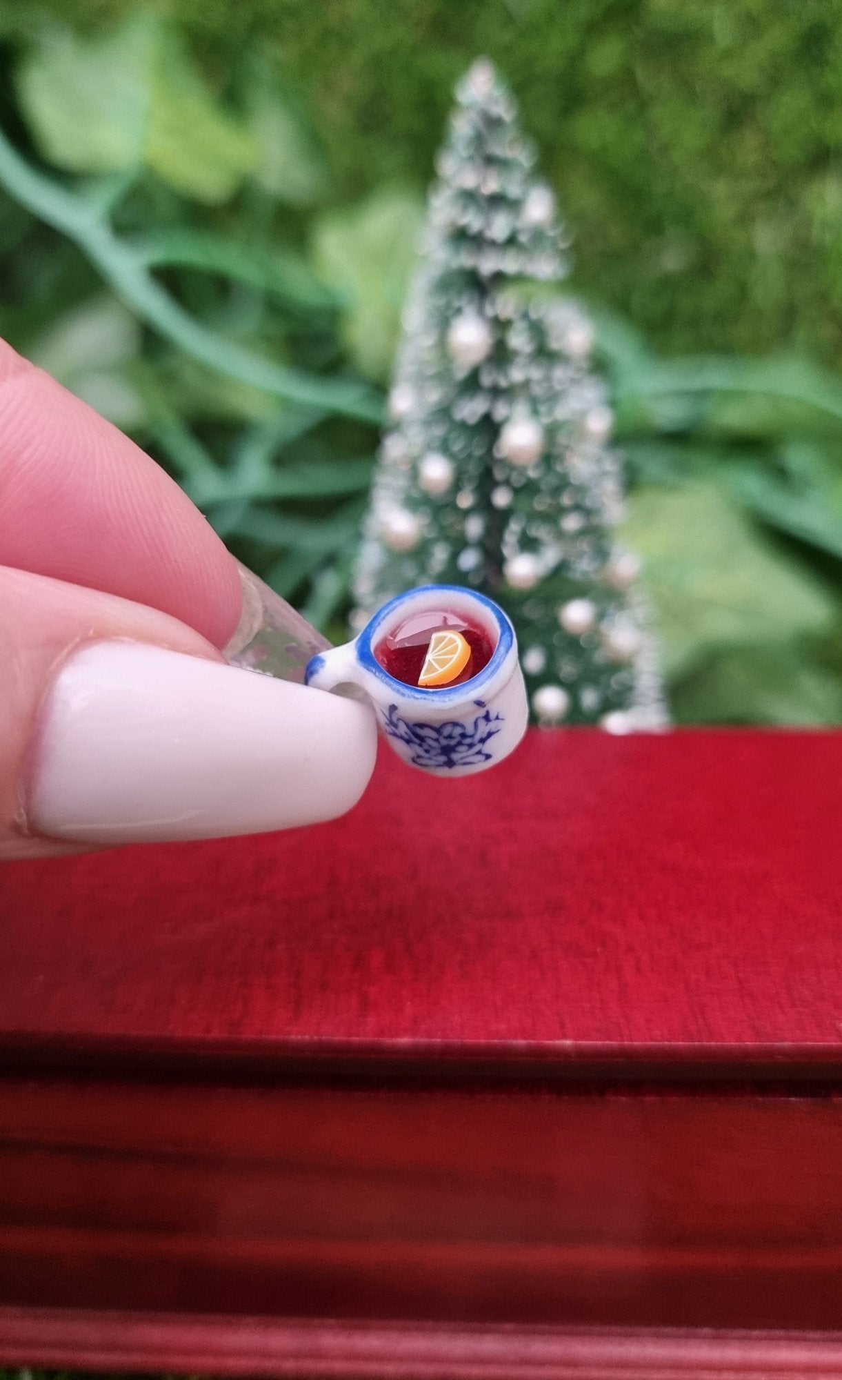 A 1:12 scale miniature mug is held between long fingernails. The tiny white and blue porcelain mug contains red liquid (mulled wine), with a slice of orange floating on the surface. The mug has an ornate pattern. Shown against a Christmas tree