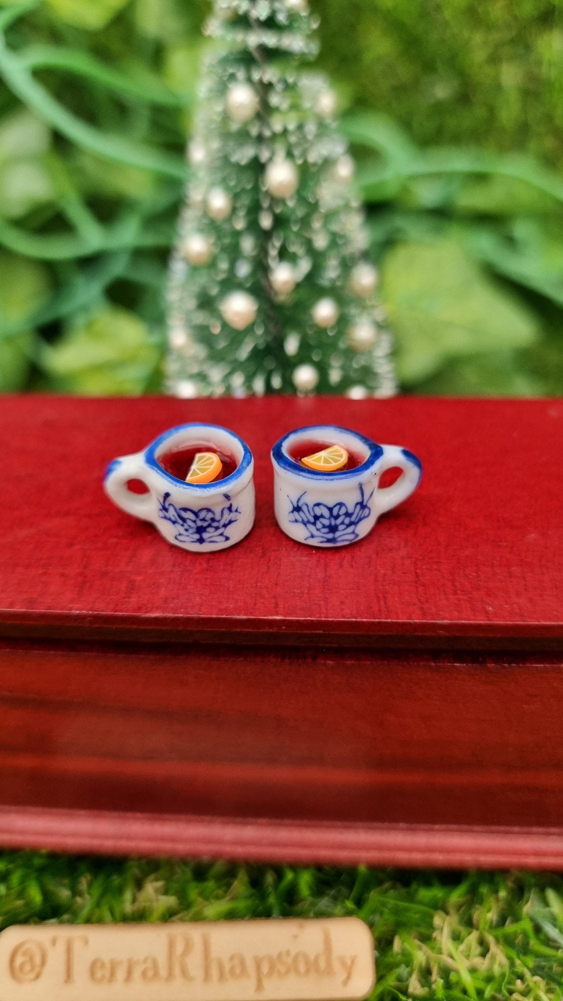 2 1:12 scale miniature mugs sit on a mini chest. The tiny white and blue porcelain mugs contain red liquid (mulled wine), with a slice of orange floating on the surface. The mug has an ornate pattern. @TerraRhapsody is written on wood
