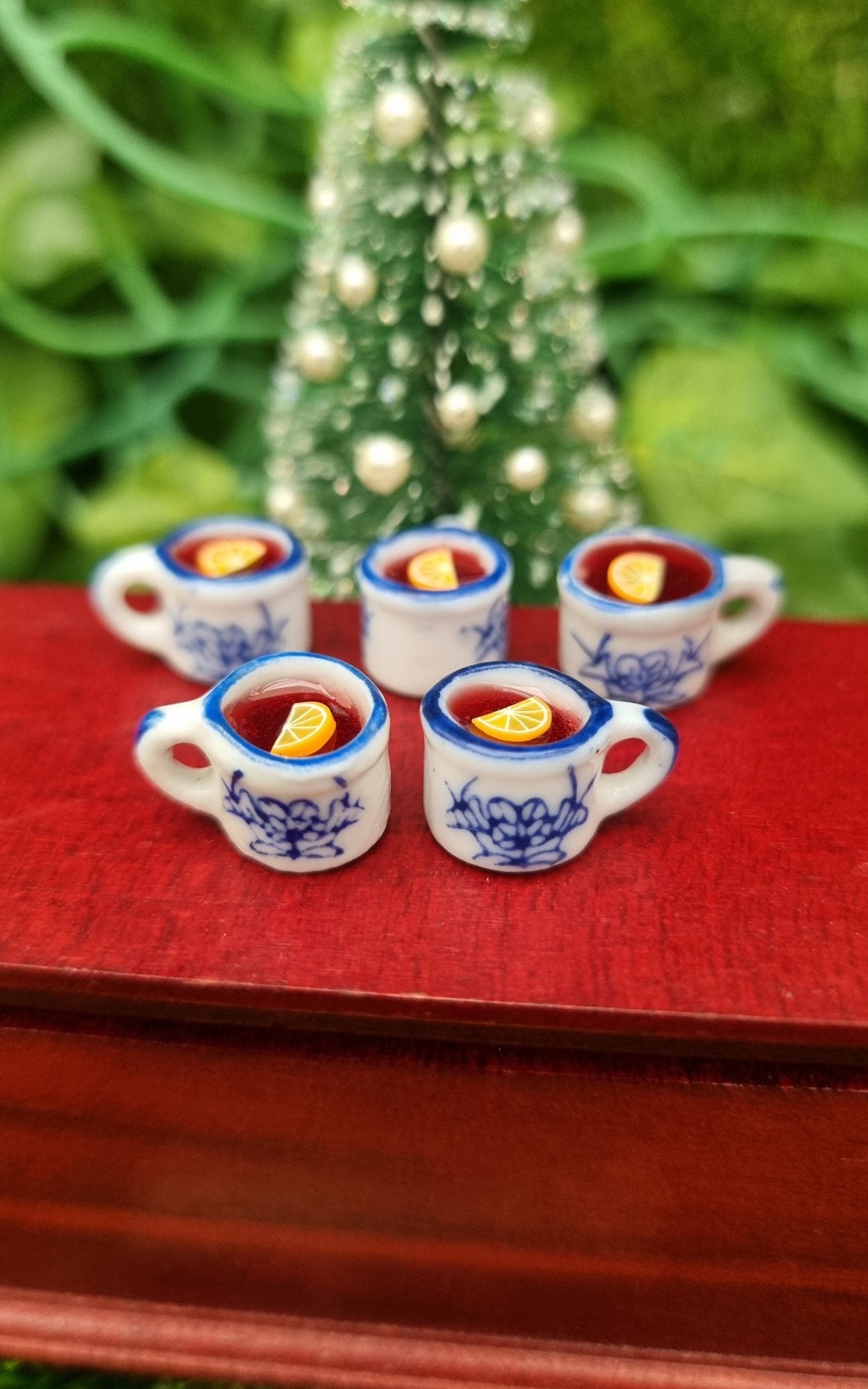 5 1:12 scale miniature mugs sit on a mini chest. The tiny white and blue porcelain mugs contain red liquid (mulled wine), with a slice of orange floating on the surface. The mugs have an ornate pattern. Shown against a Christmas tree
