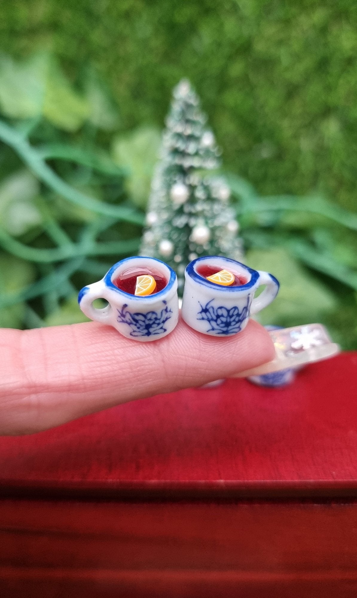 2 1:12 scale miniature mugs are held on the end of a finger. The tiny white and blue porcelain mugs contain red liquid (mulled wine), with a slice of orange floating on the surface. The mugs have an ornate pattern. Shown against a Christmas tree