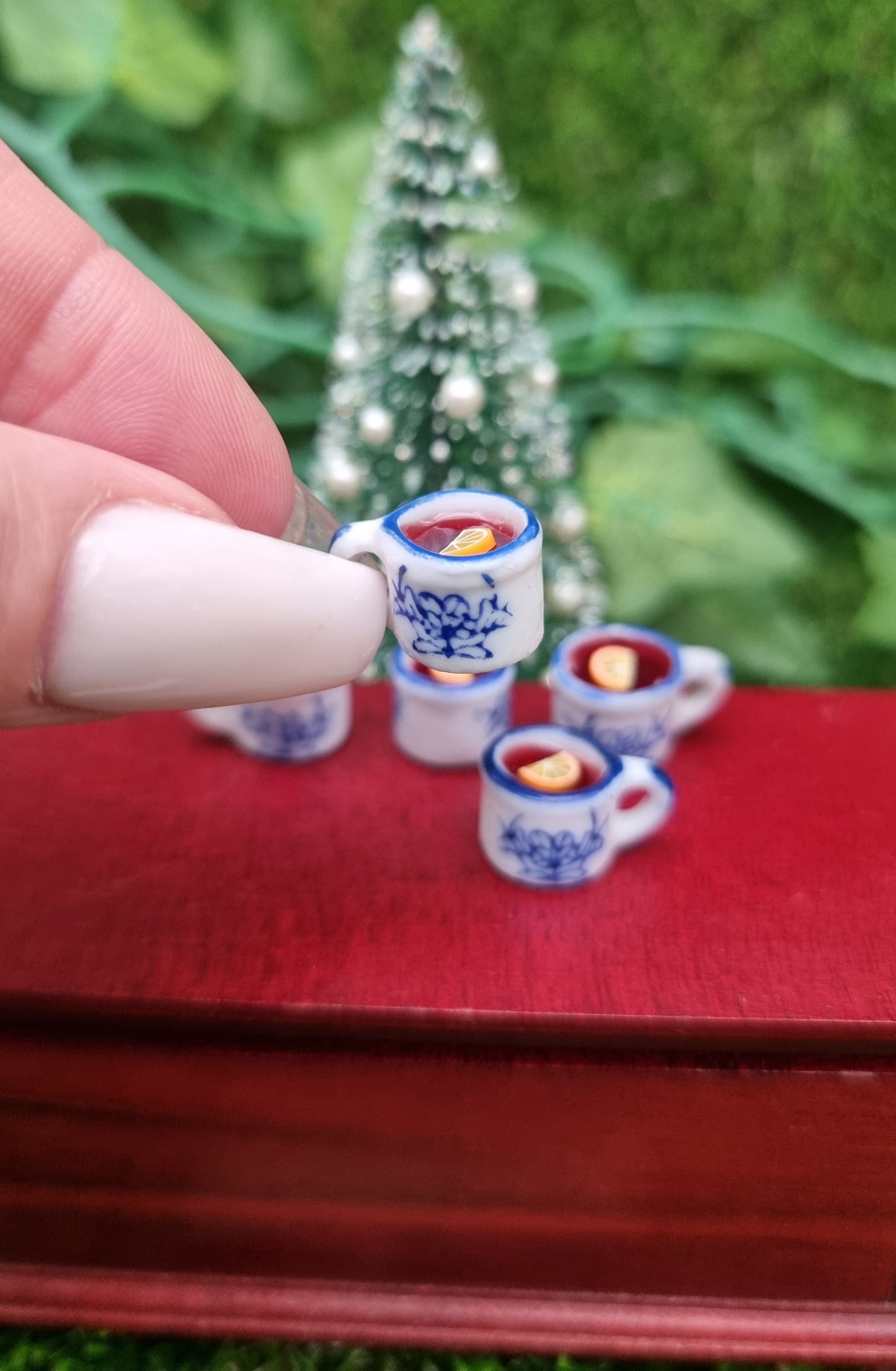 A 1:12 scale miniature mug is held between long fingernails. The tiny white and blue porcelain mug contains red liquid (mulled wine), with a slice of orange floating on the surface. The mug has an ornate pattern. Christmas tree and more mugs behind