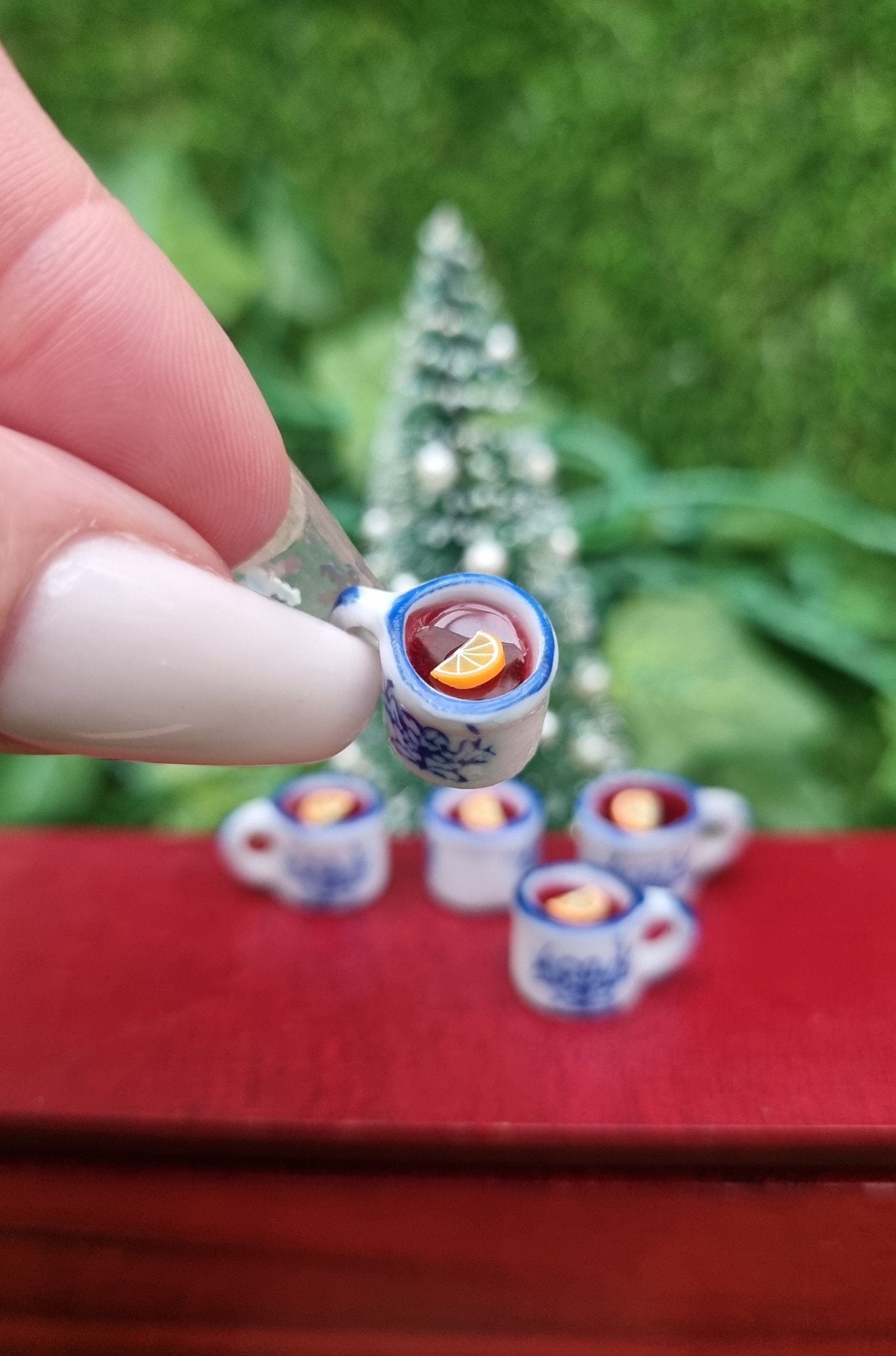A 1:12 scale miniature mug is held between long fingernails. The tiny white and blue porcelain mug contains red liquid (mulled wine), with a slice of orange floating on the surface. The mug has an ornate pattern. Christmas tree and more mugs behind