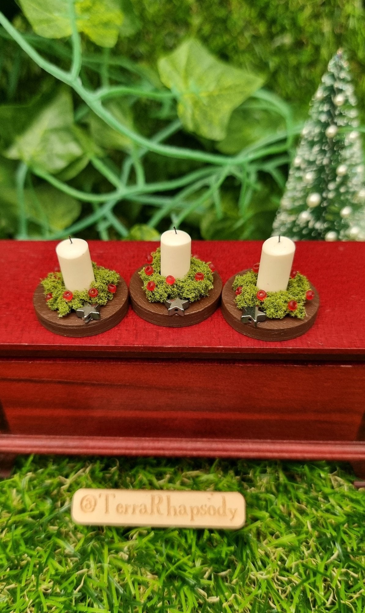 1:12 scale miniature candle centrepieces sit on round brown plates on top of a wooden chest. There are 3 plates each with an ivory candle, green foliage, red berries and silver star. @Terra Rhapsody is written on wood in front.