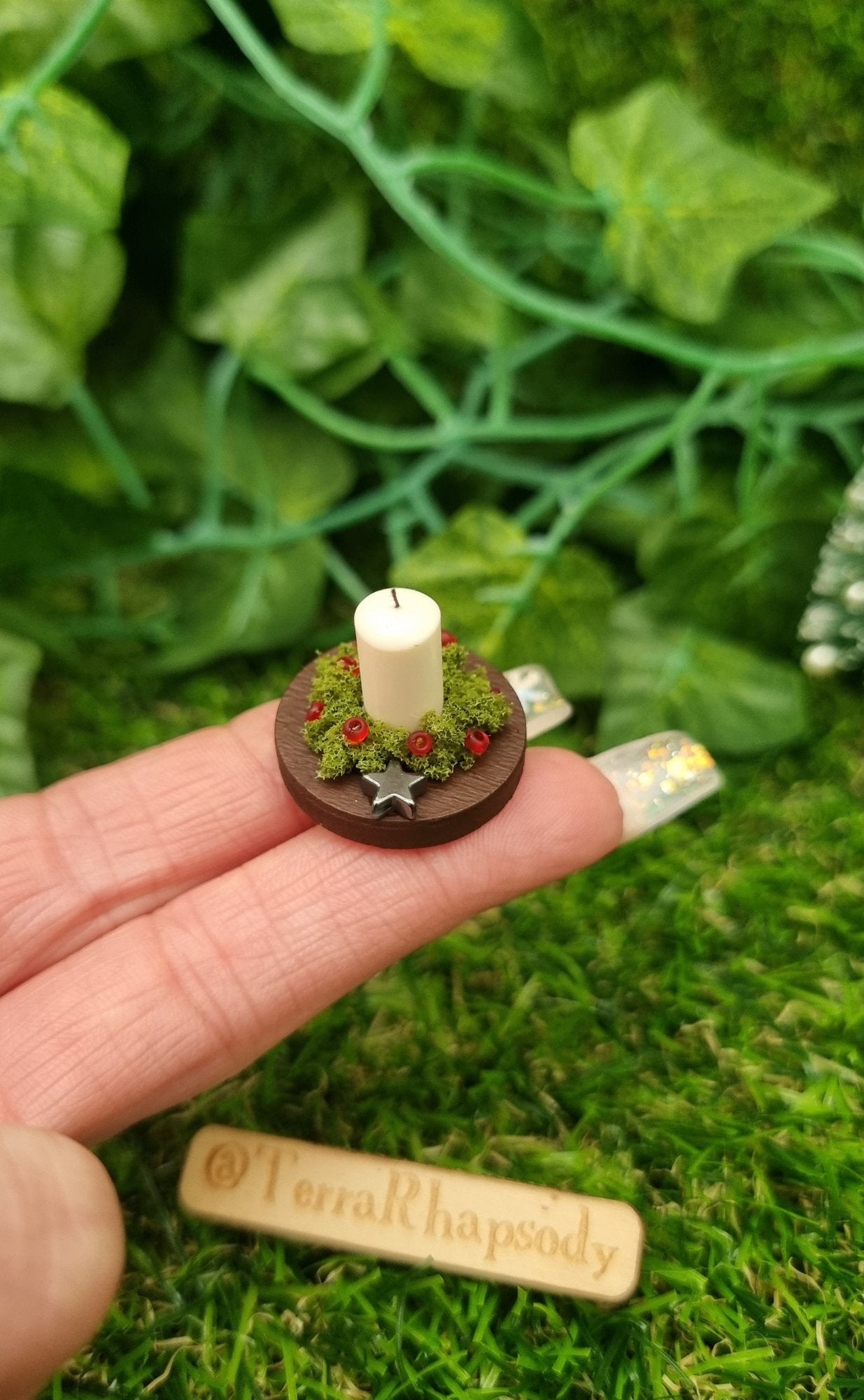 1:12 scale miniature candle centrepiece sits on a round brown plate on fingertips in front of a wooden chest. It has an ivory candle, green foliage, red berries and silver star.@TerraRhapsody is written on wood in the background