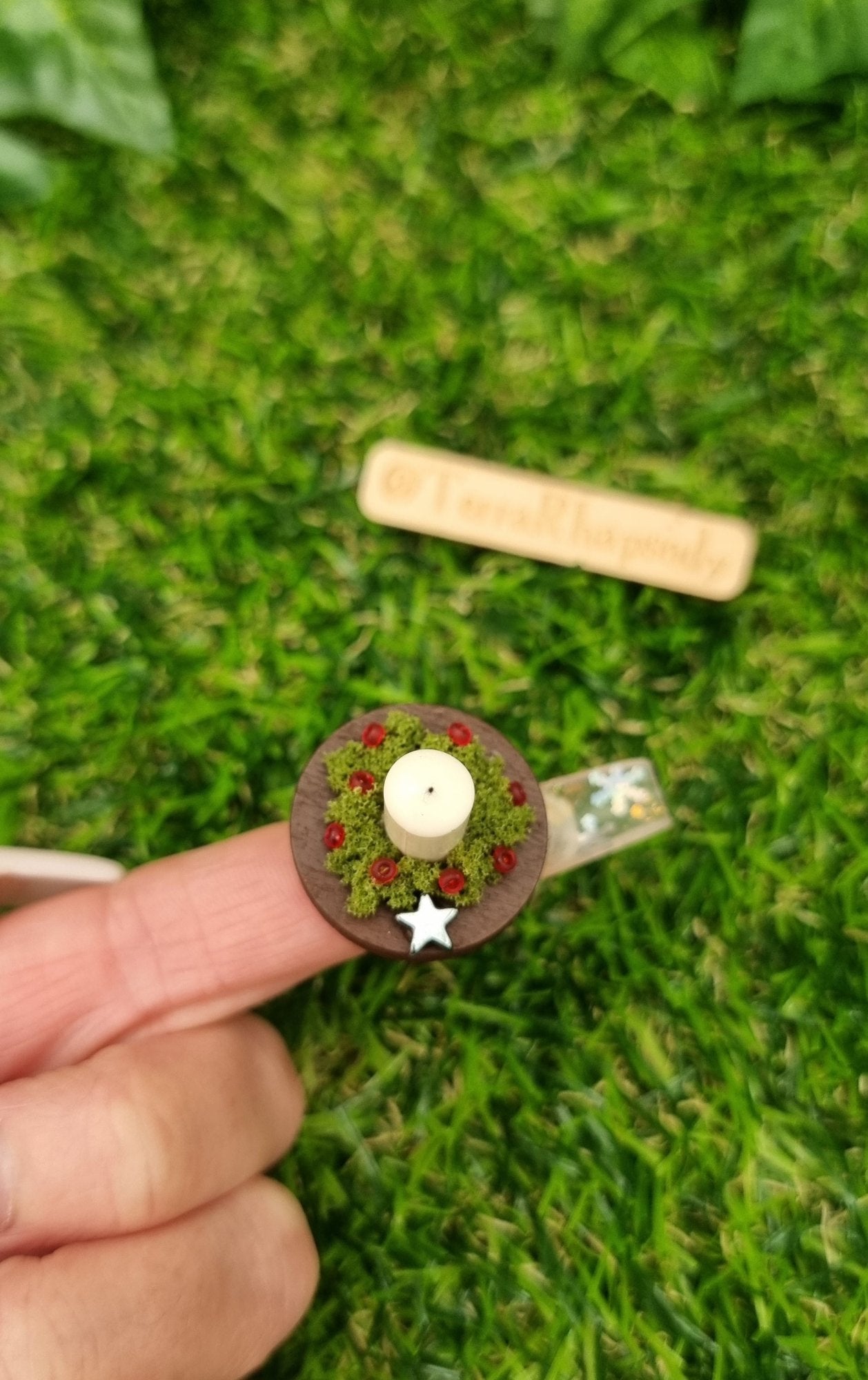 1:12 scale miniature candle centrepiece sits on a round brown plate on a fingertip in front of a wooden chest. It has an ivory candle, green foliage, red berries and silver star. Shown from above @TerraRhapsody is written on wood in the background