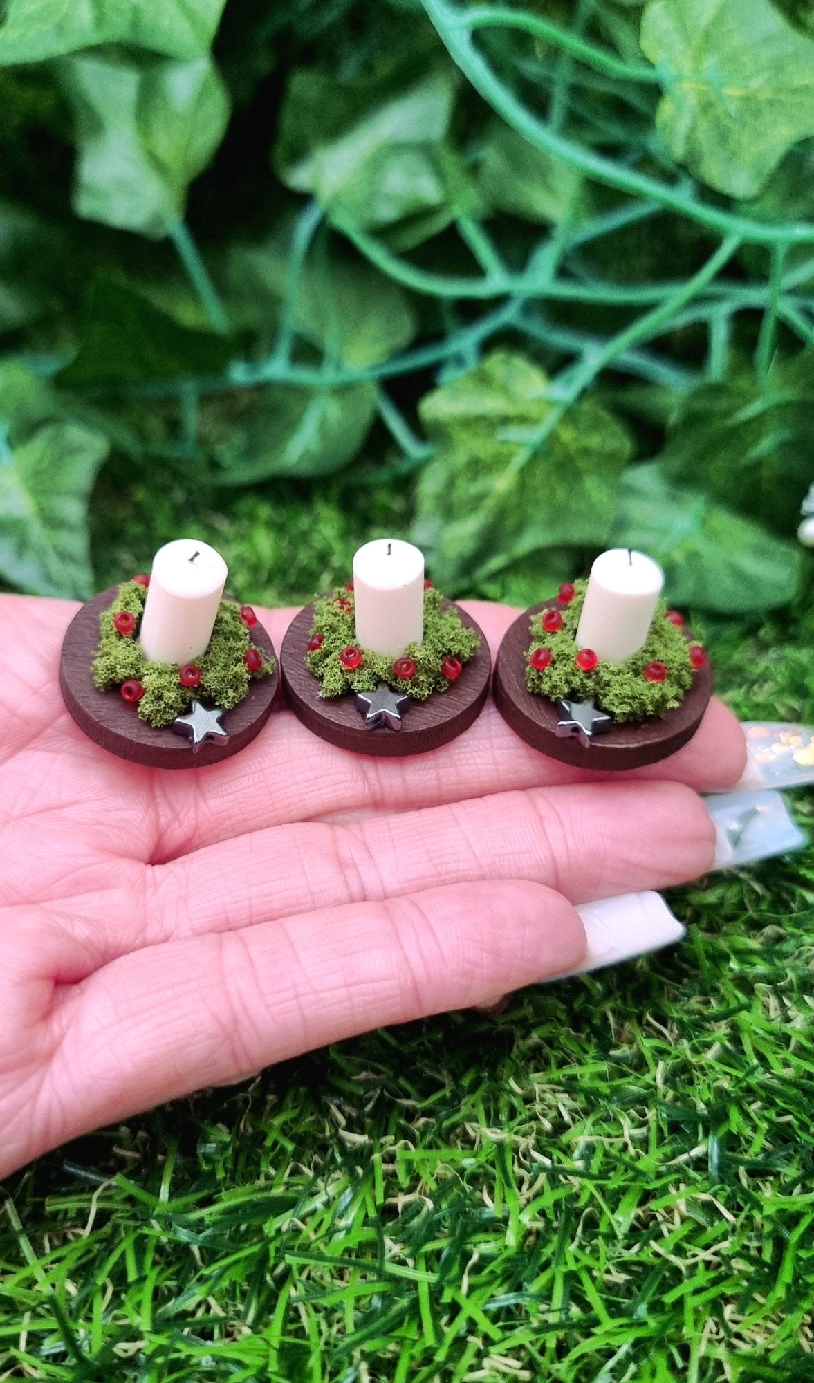1:12 scale miniature candle centrepieces sit on round brown plates on top of a wooden chest. There are 3 plates each with an ivory candle, green foliage, red berries and silver star. @Terra Rhapsody is written on wood in front.