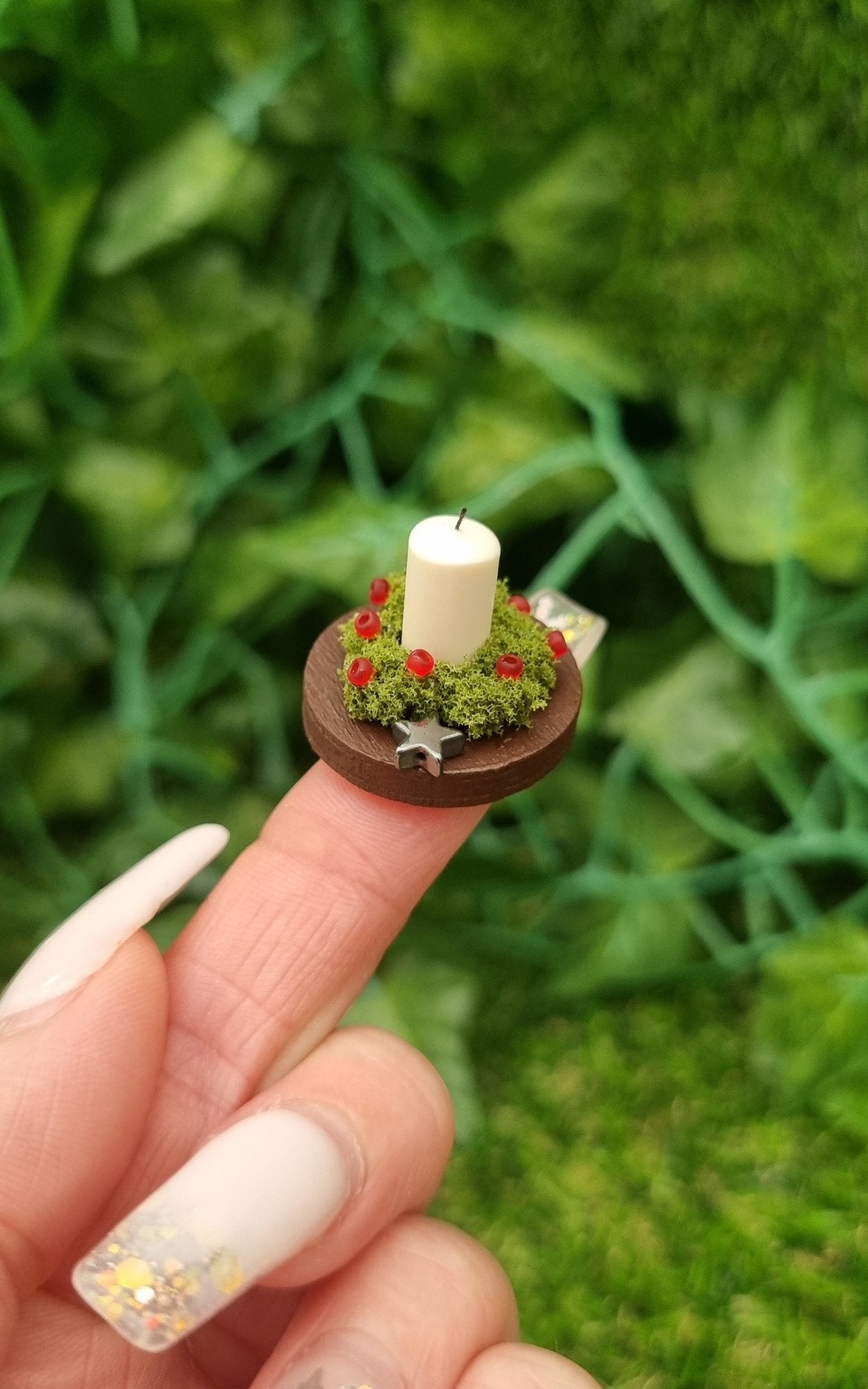 1:12 scale miniature candle centrepiece sits on a round brown plate on a fingertip in front of a wooden chest. It has an ivory candle, green foliage, red berries and silver star.@TerraRhapsody is written on wood in the background