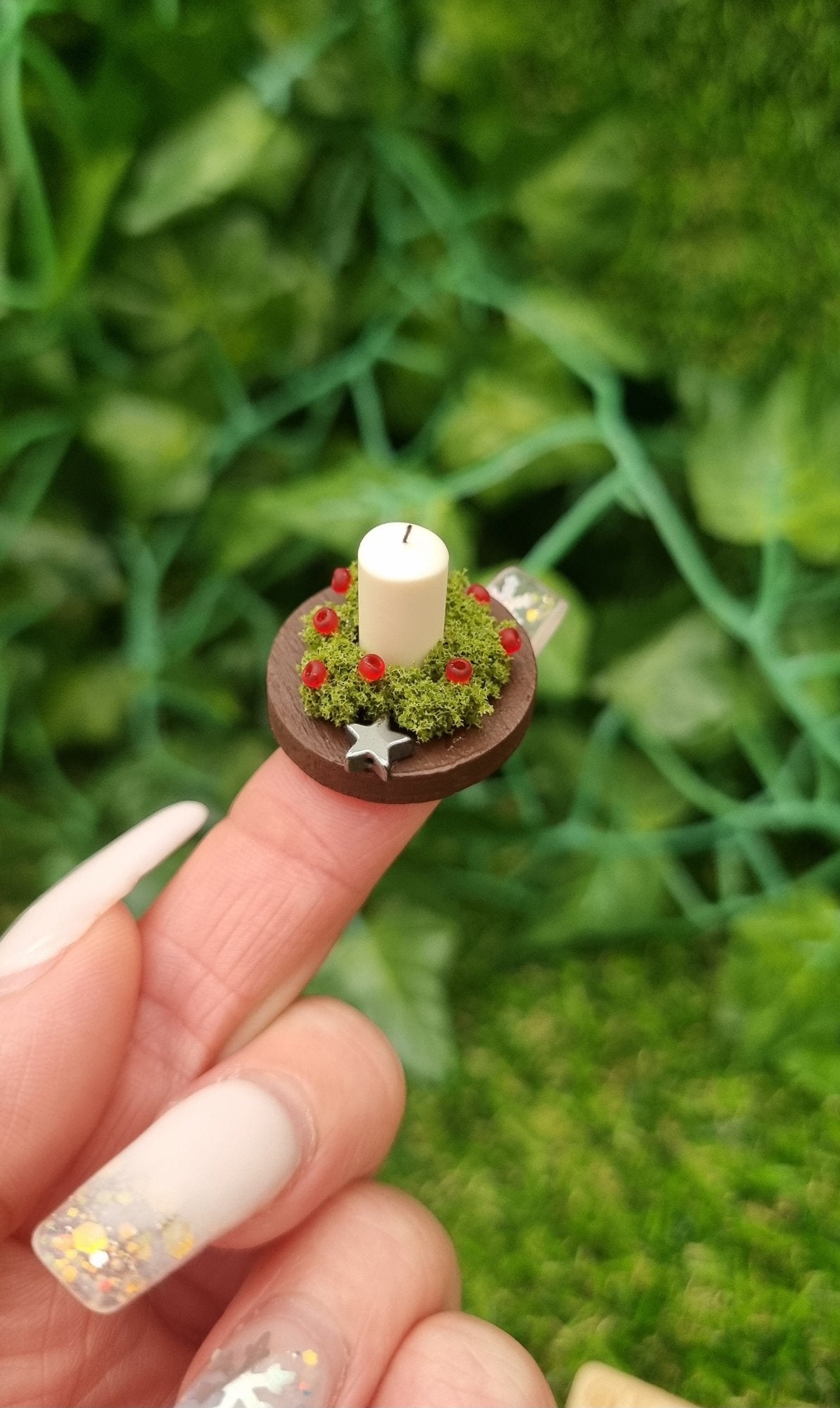 1:12 scale miniature candle centrepiece sits on a round brown plate on a fingertip in front of a wooden chest. It has an ivory candle, green foliage, red berries and silver star.@TerraRhapsody is written on wood in the background