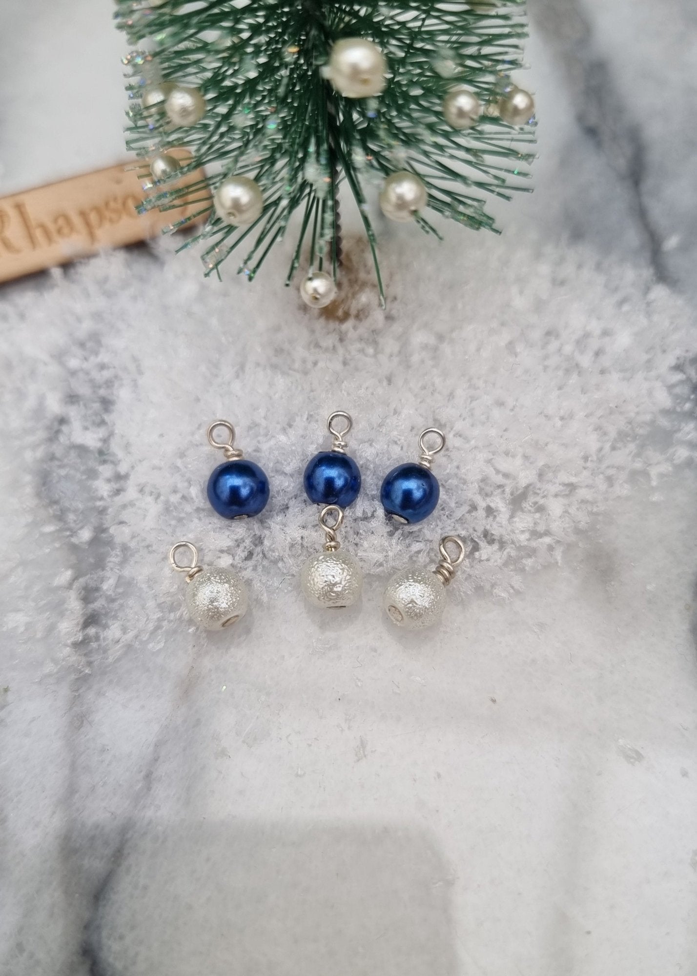 6 Miniature 1:12 Christmas baubles are displayed on snow at the foot of a miniature Christmas tree. Three baubles are round and pearly blue and three are textured white in colour. @TerraRhapsody is written on wood in the background