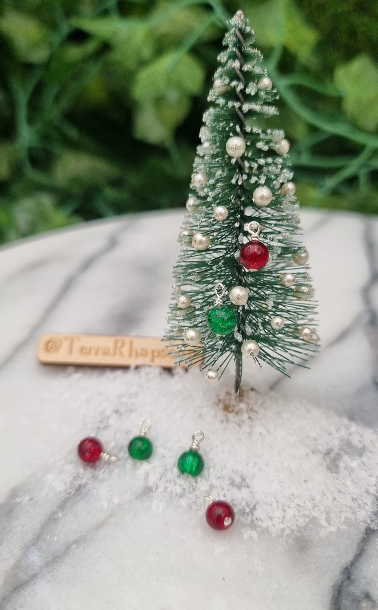 Miniature 1:12 Christmas baubles are displayed on a tiny Christmas tree. The baubles are round crackle glass in bright red and green festive colours. More baubles sit in snow at the foot of the tree. @TerraRhapsody is written on wood in the background