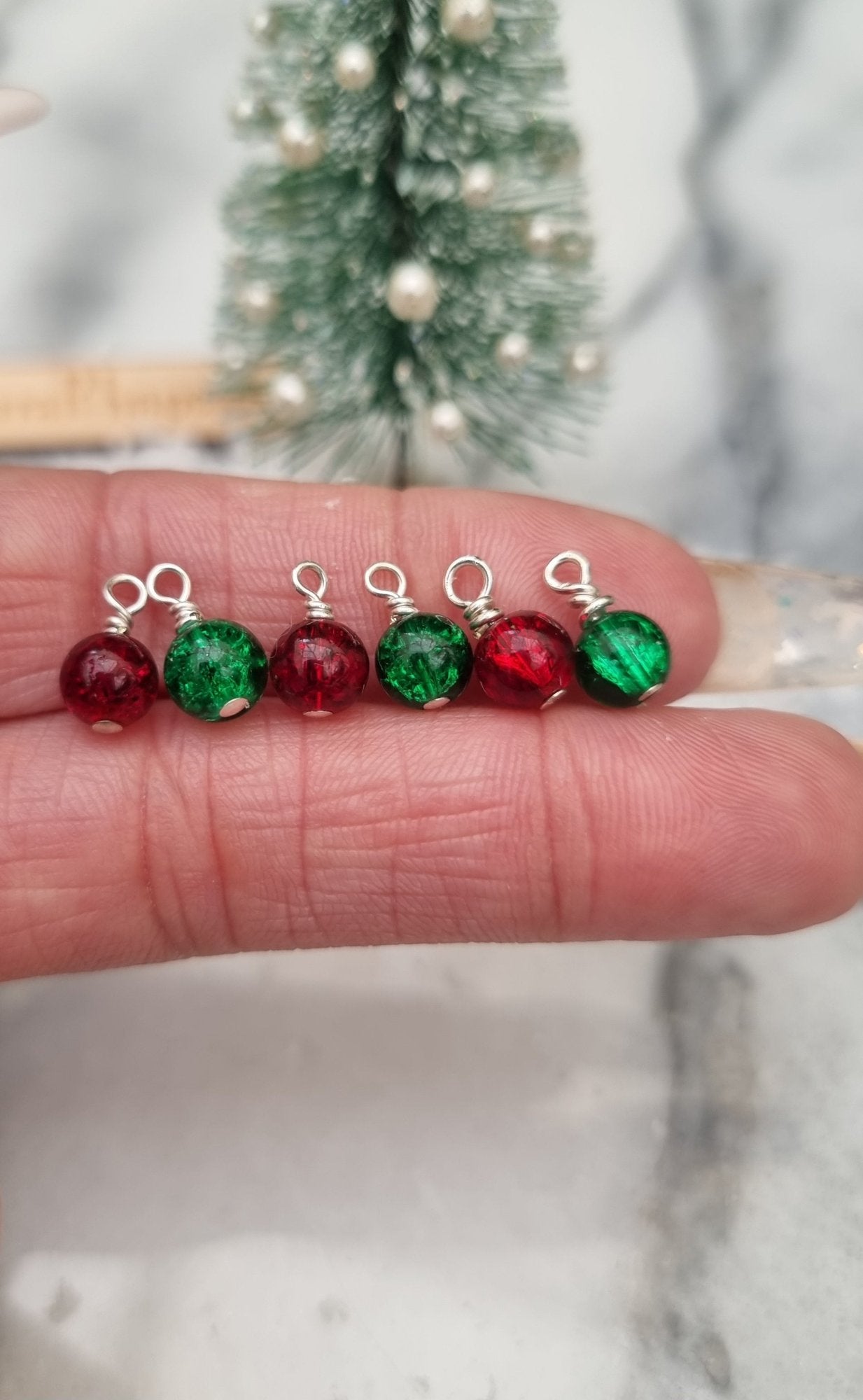 6 Miniature 1:12 Christmas baubles are displayed on a hand front of a miniature Christmas tree. The baubles are round crackle glass in bright red and green colours. @TerraRhapsody is written on wood in the background